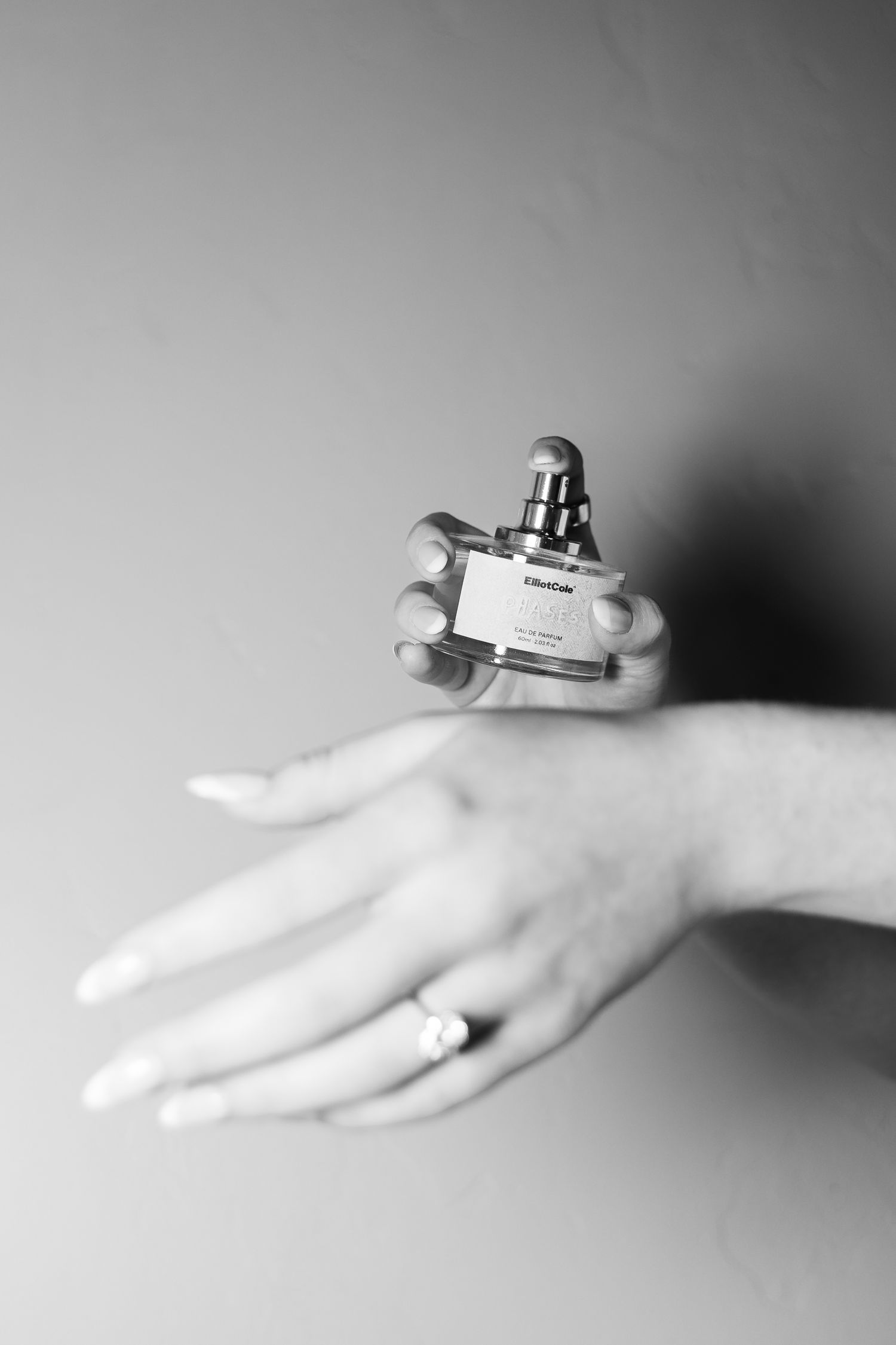 Black and white photo of a hand holding a small perfume bottle while another hand with a ring rests below.