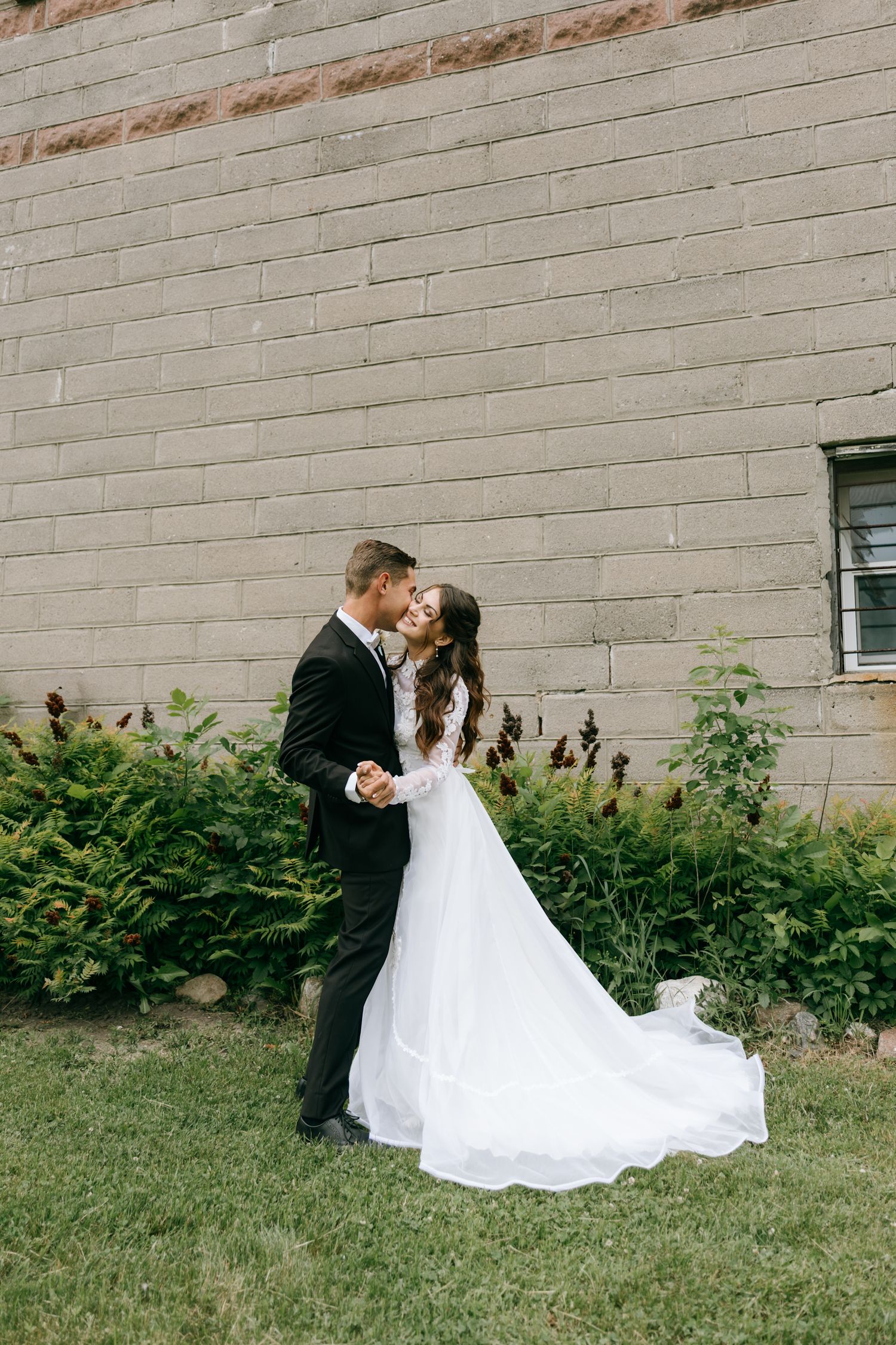A bride and groom share a romantic kiss outdoors, with the bride's white gown flowing gracefully on green grass.