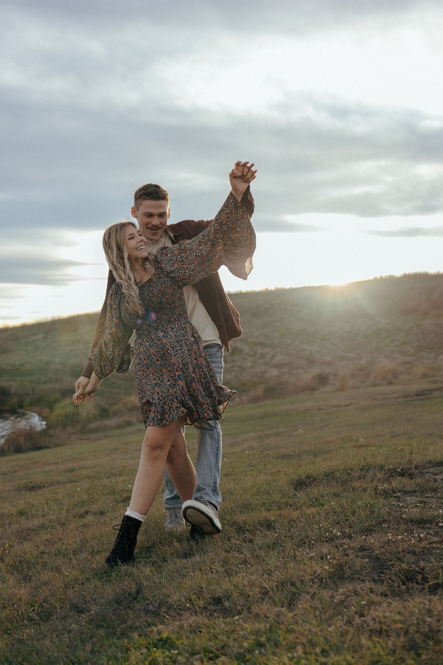 Playful dancing scene on a grassy hillside during sunset with cloudy skies.