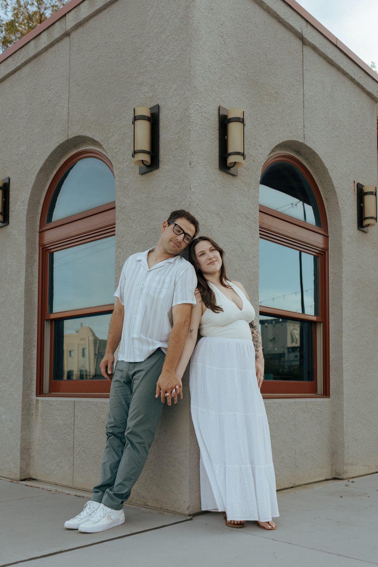 Two people lean against a building exterior with arched windows in casual poses.