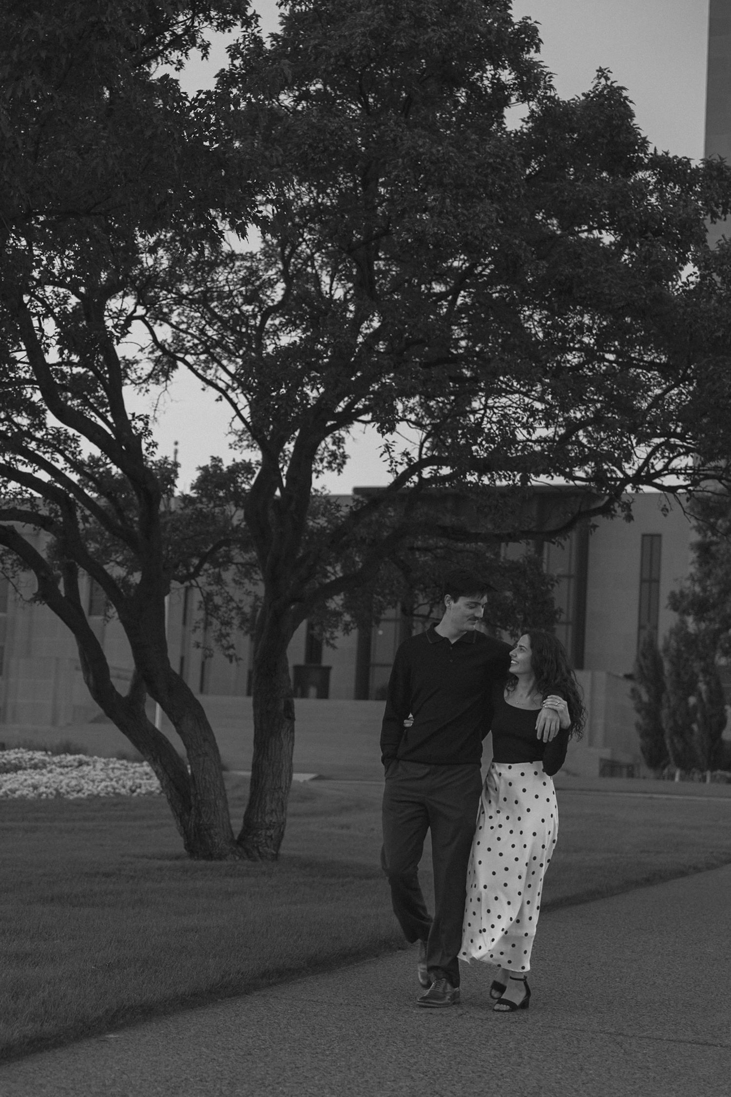 A couple shares a romantic moment outdoors under large trees in black and white.