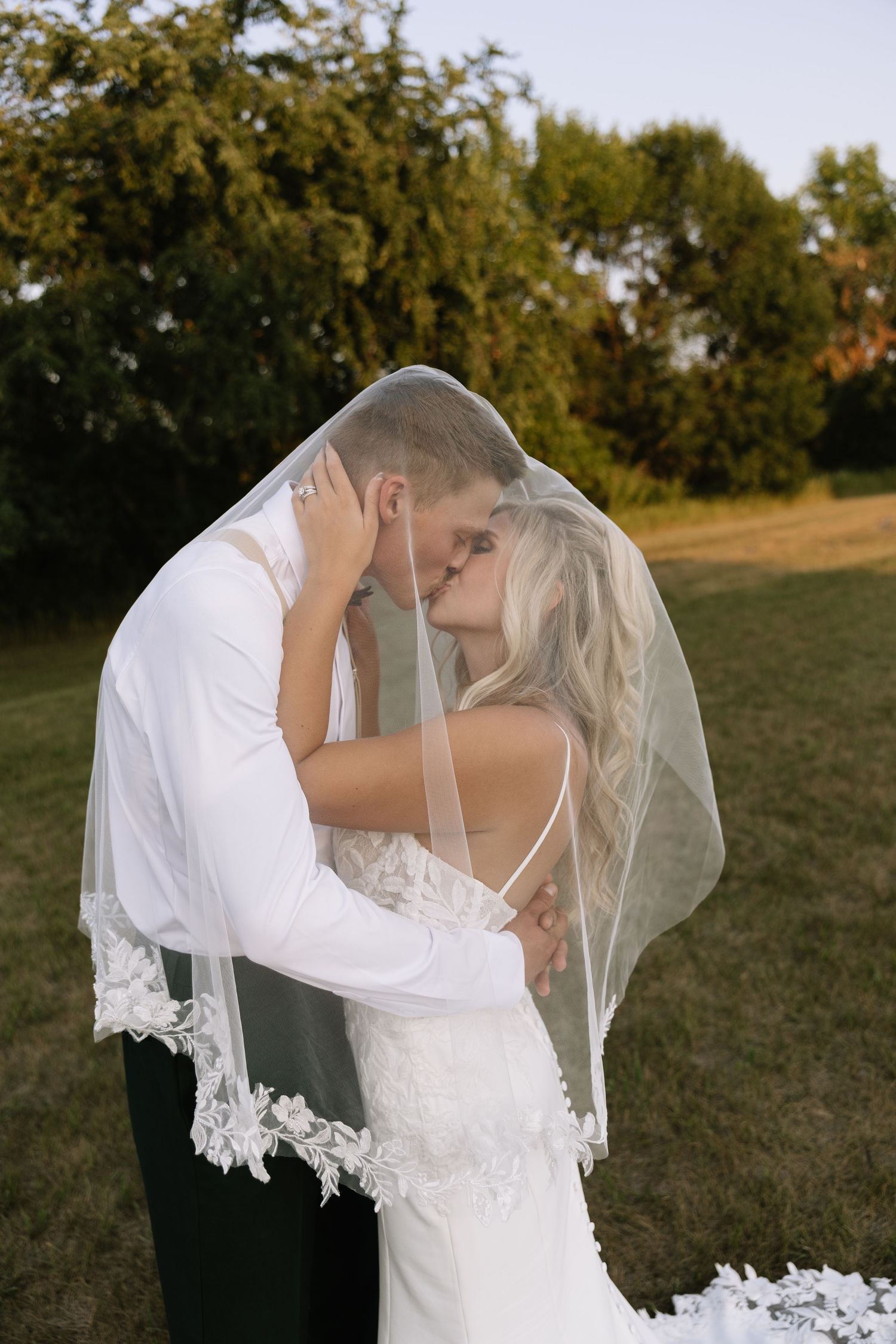 Bride and groom sharing a romantic kiss under a lace-trimmed veil outdoors on their wedding day.