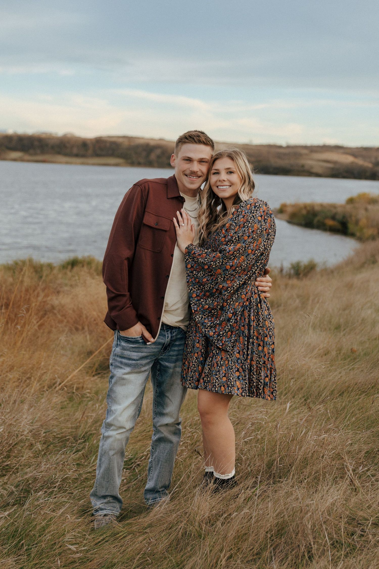 Couple in casual fall outfits posing together by lake with rolling hills.