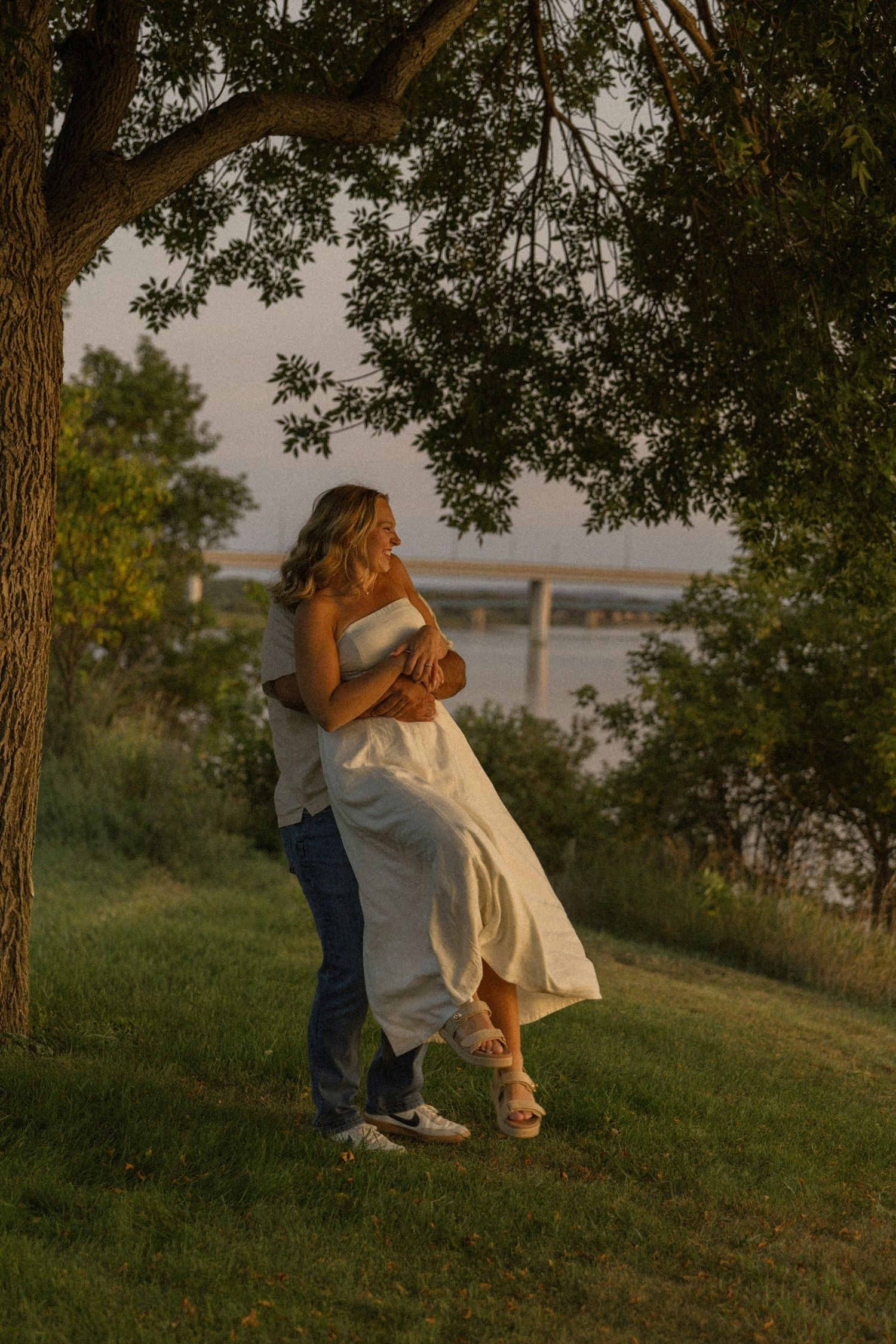 A couple dances playfully on a grassy hillside near a lake during golden hour.