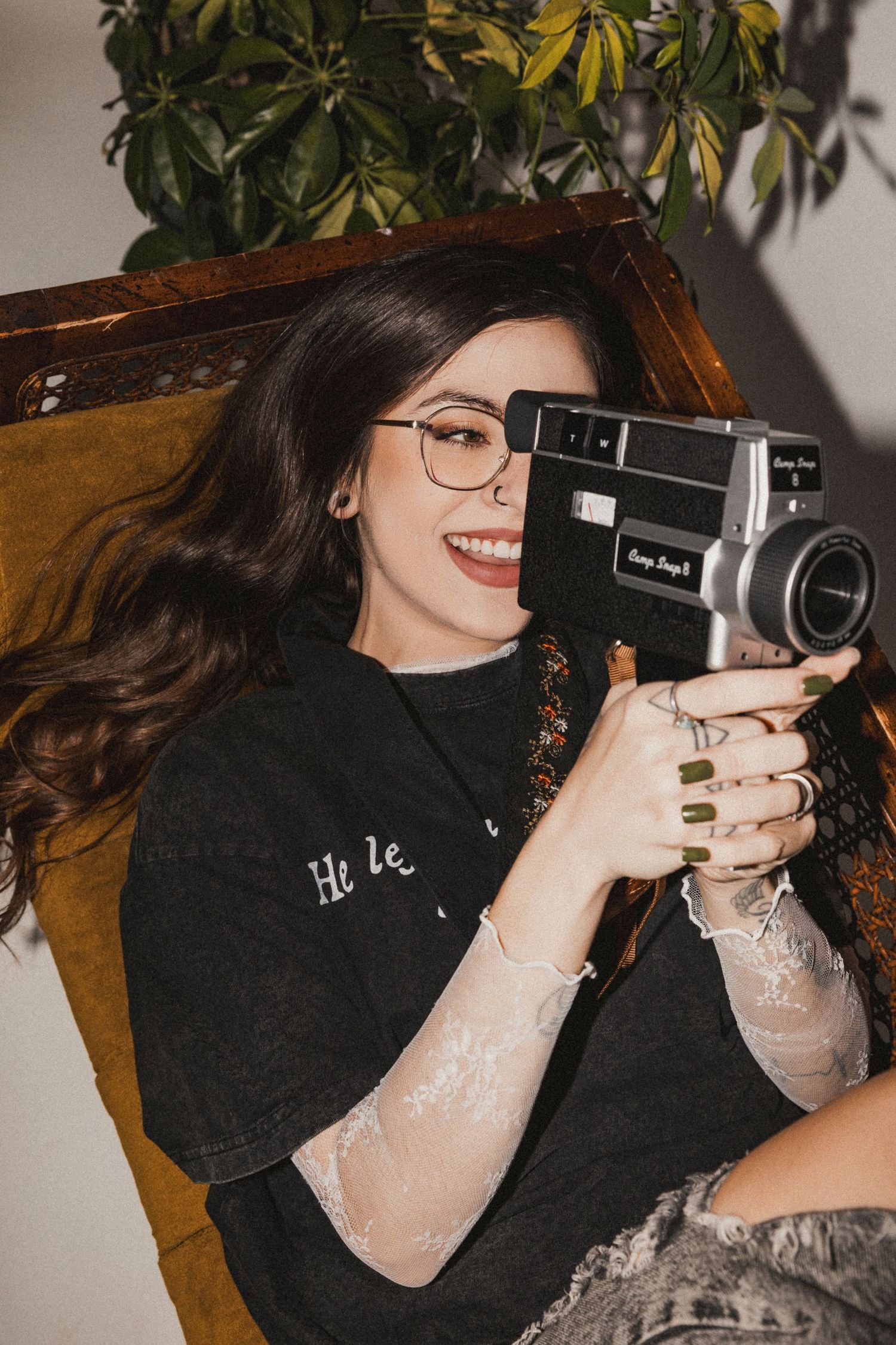 Smiling woman with glasses holding a vintage Super 8 camera, wearing a black tee, seated in a wooden chair near green plants.