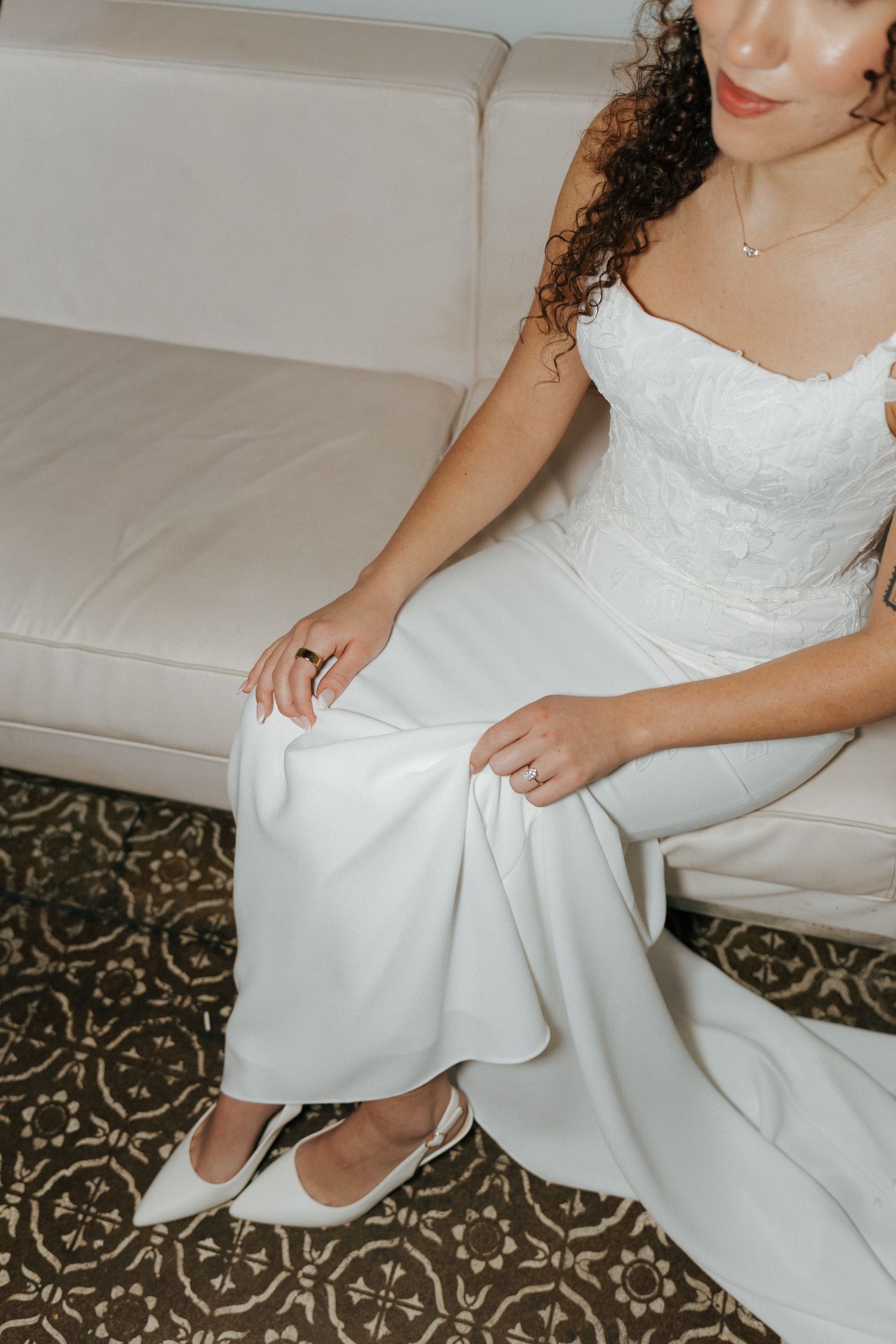 Bride in white lace wedding dress and white flats seated on ivory sofa, adjusting her gown skirt.