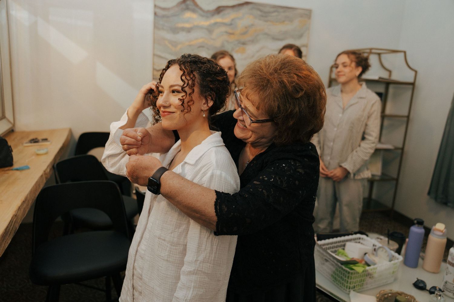 Older woman helping bride get dressed in bridal suite while guests watch in background.