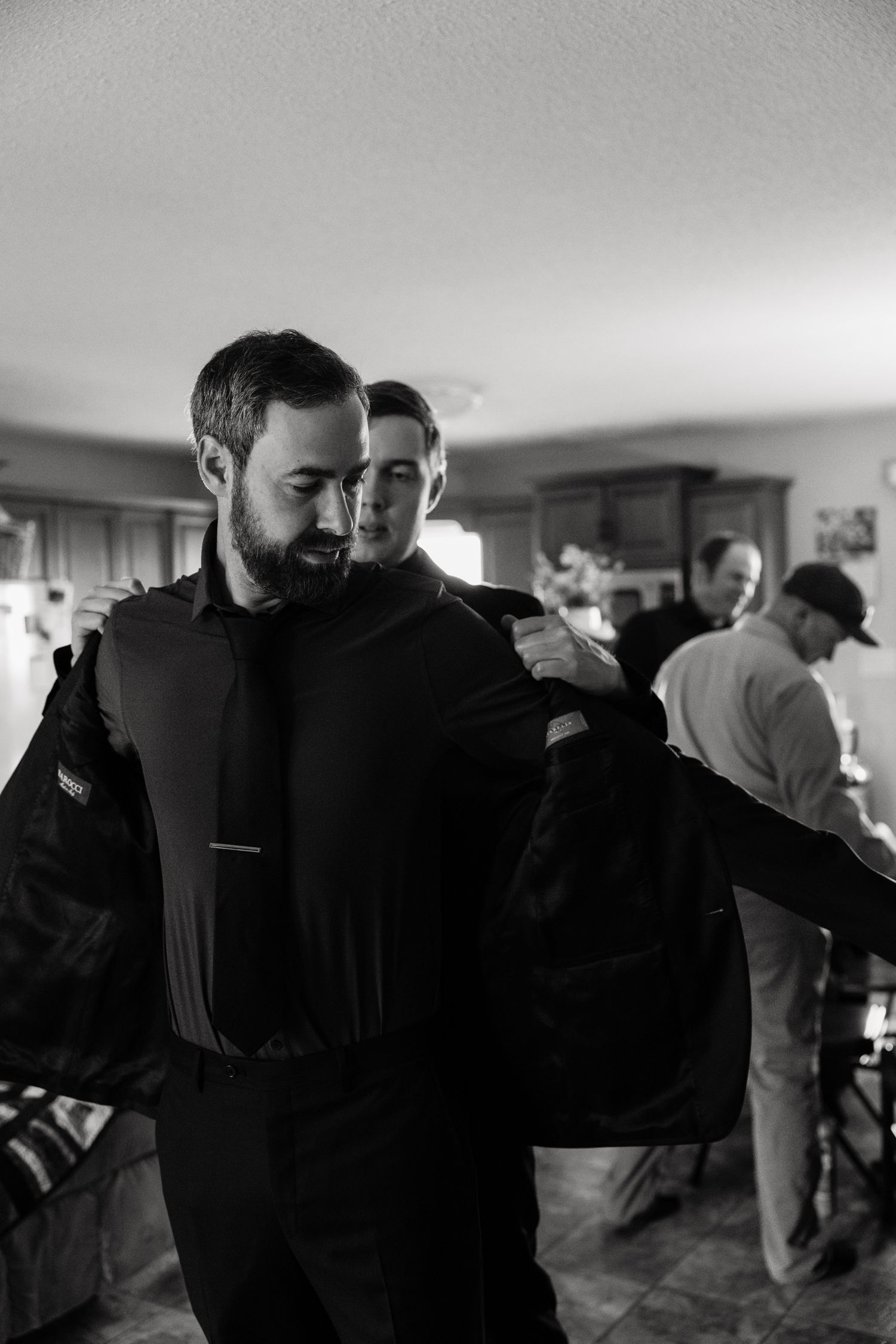 Black and white photo of a bearded man being helped into a suit jacket by a friend indoors.