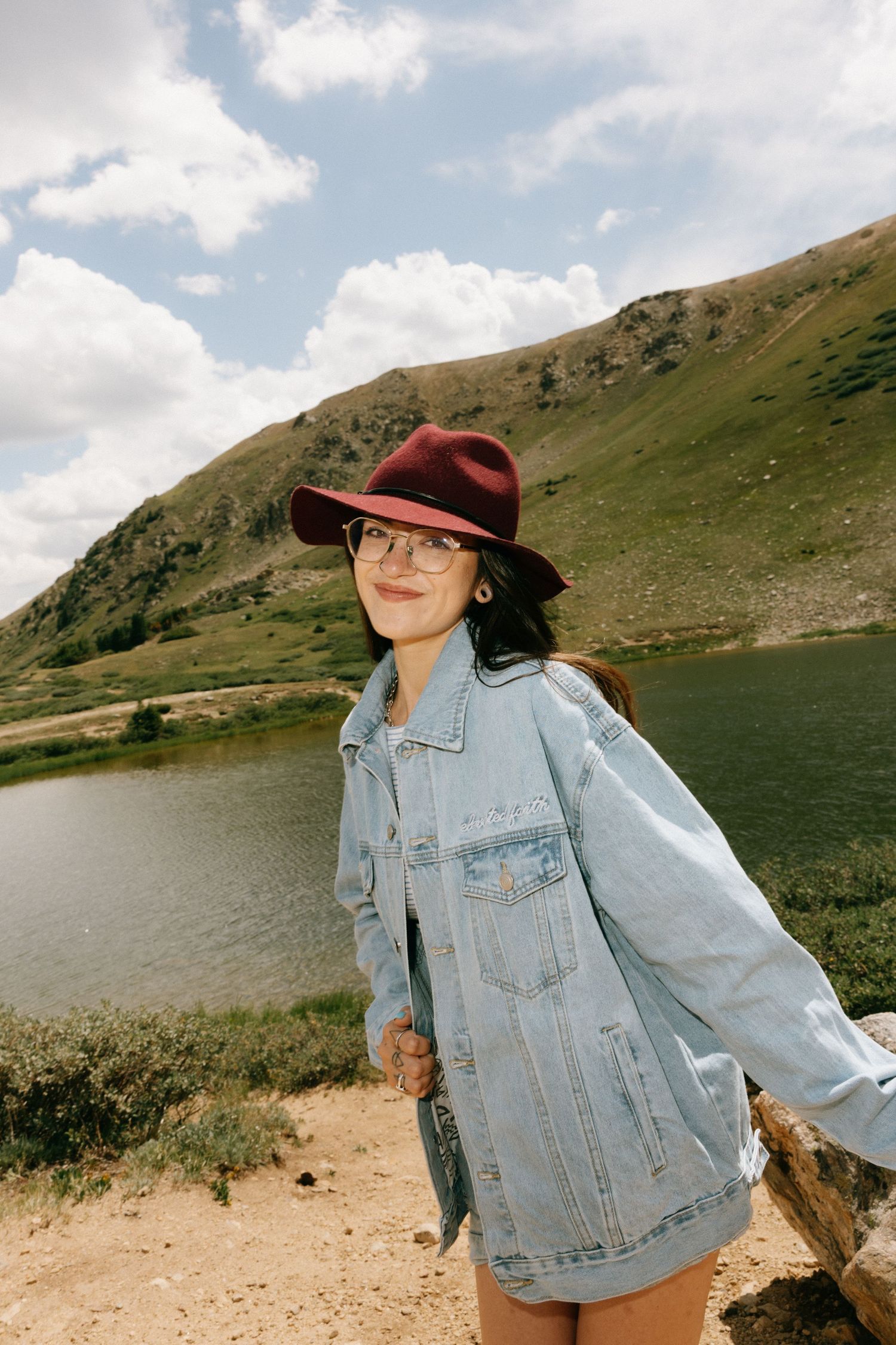 Woman in denim jacket and burgundy hat smiling by a mountain lake under cloudy skies.