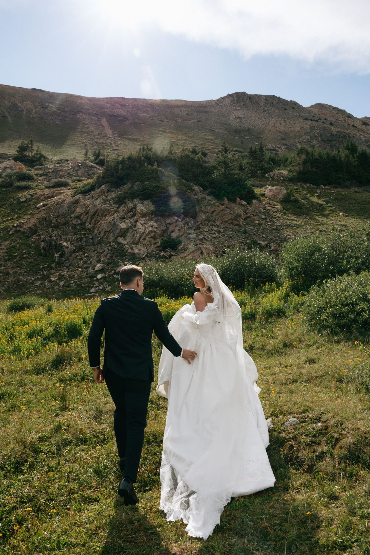 Bride and groom walking hand in hand through a lush green mountain landscape on their wedding day.