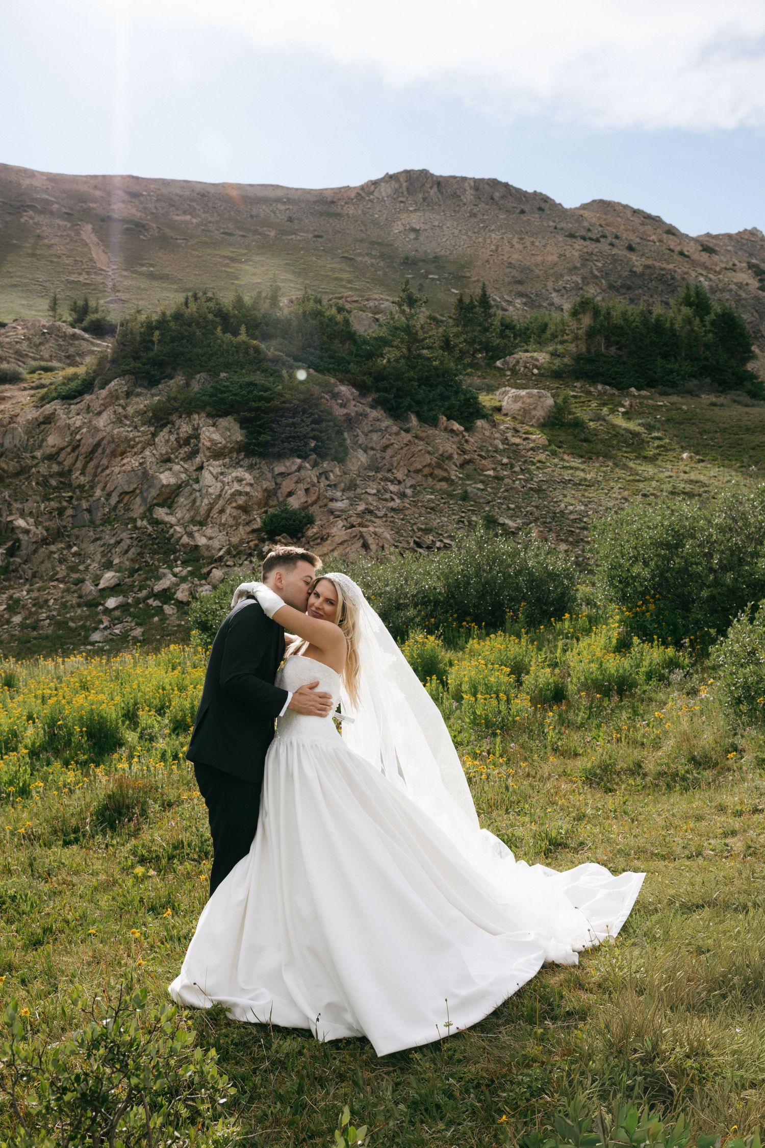 A bride and groom embrace in a mountain meadow, her white gown flowing, surrounded by wildflowers and rocky hills.