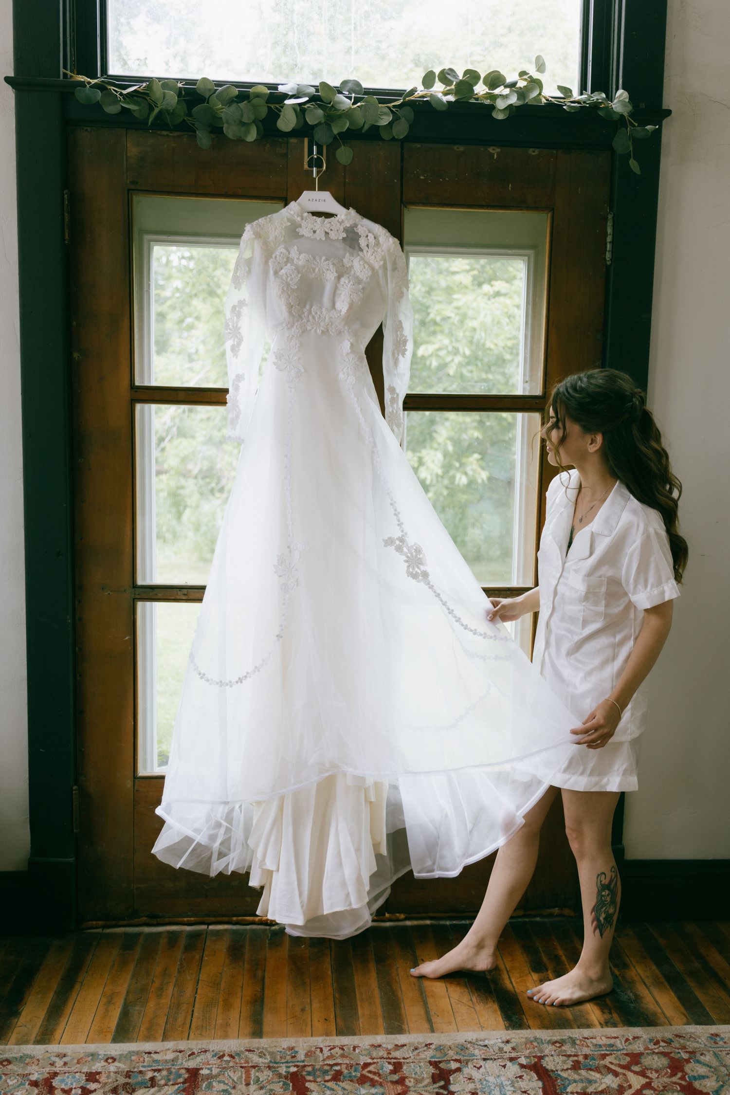 Bride admiring her white lace long-sleeve wedding gown hanging by a window in a rustic wood-paneled room.