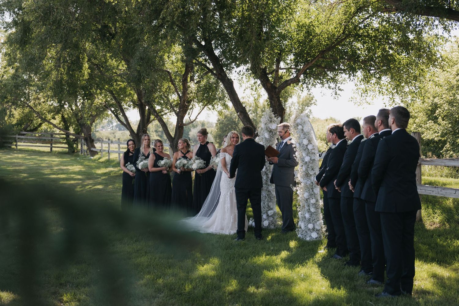 Outdoor wedding ceremony under lush green trees with bridesmaids in navy dresses and groomsmen in dark suits.