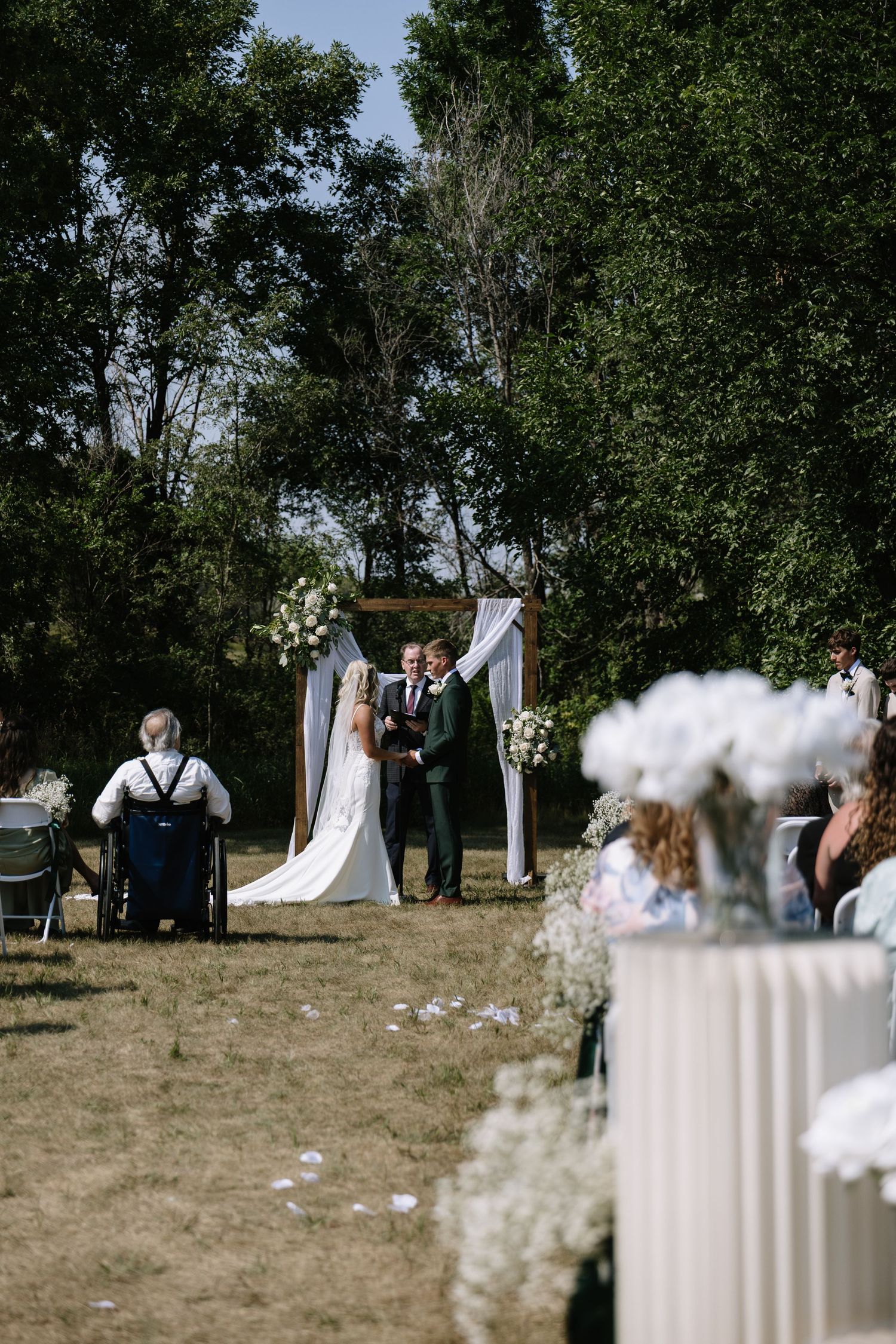 Outdoor wedding ceremony with couple at floral arch, guests seated on chairs, surrounded by lush green trees on a sunny day.