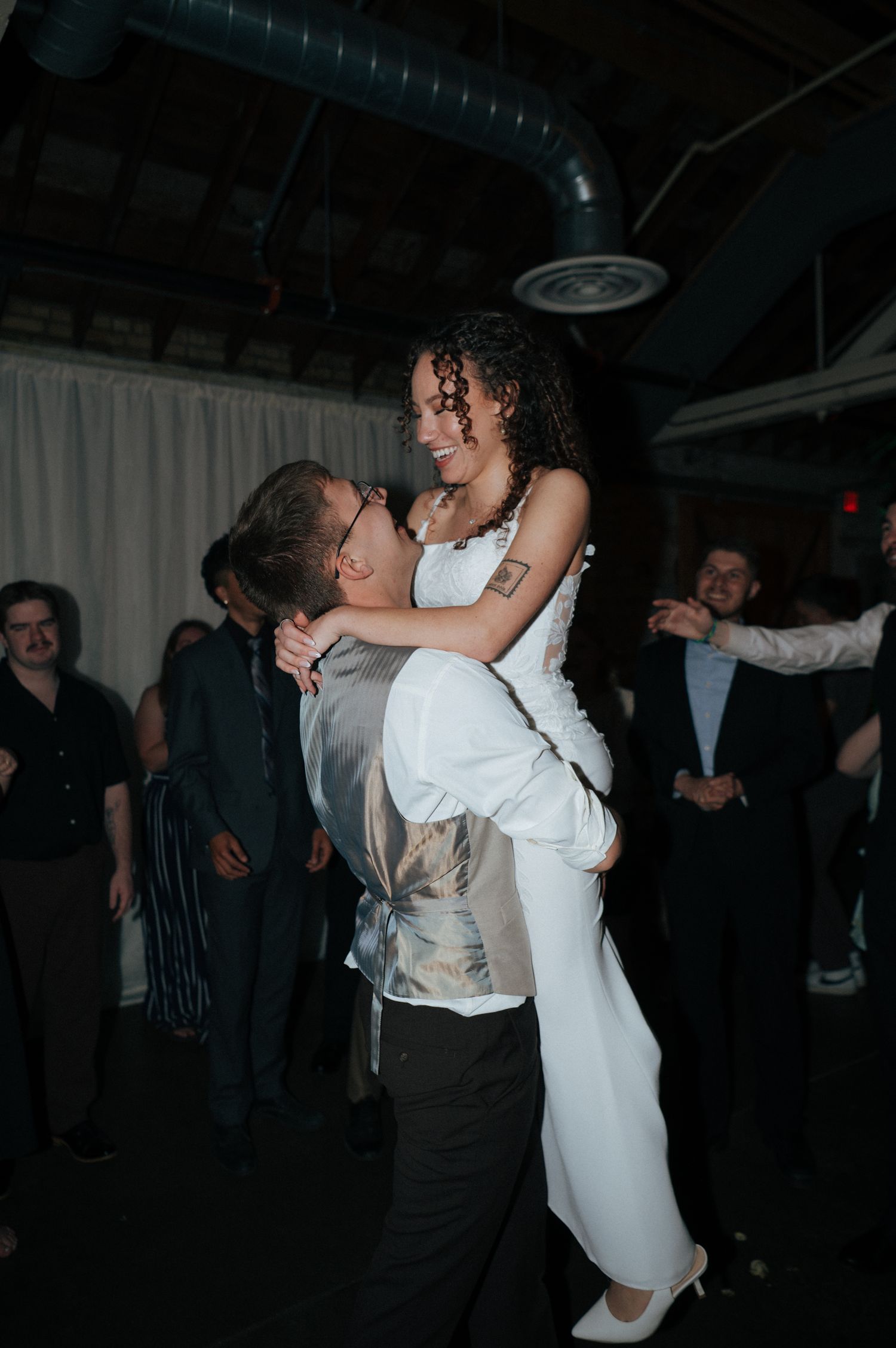Groom lifting bride in white jumpsuit during wedding reception dance floor celebration.