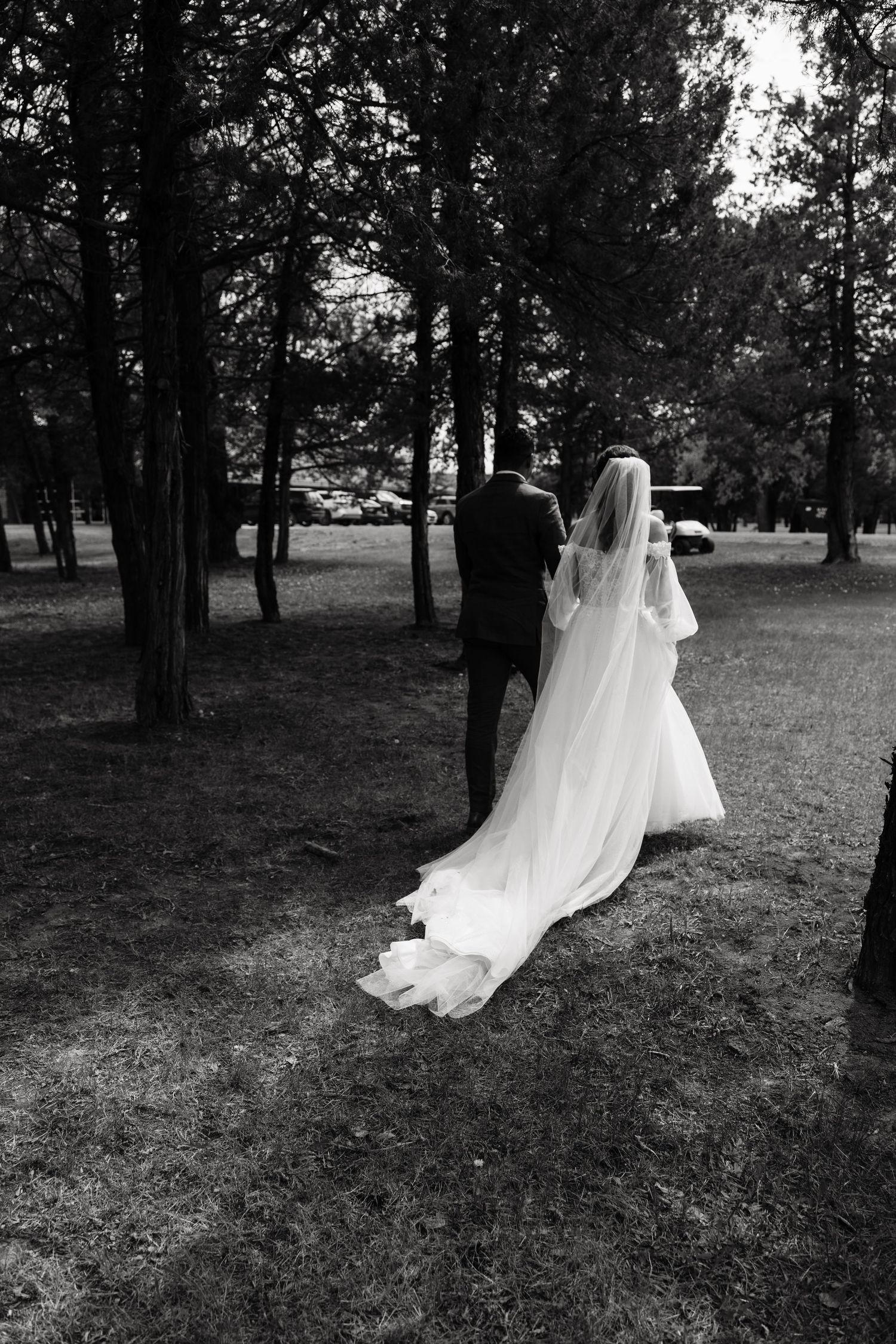 Black and white photo of bride and groom walking hand in hand through a tree-lined park on their wedding day.