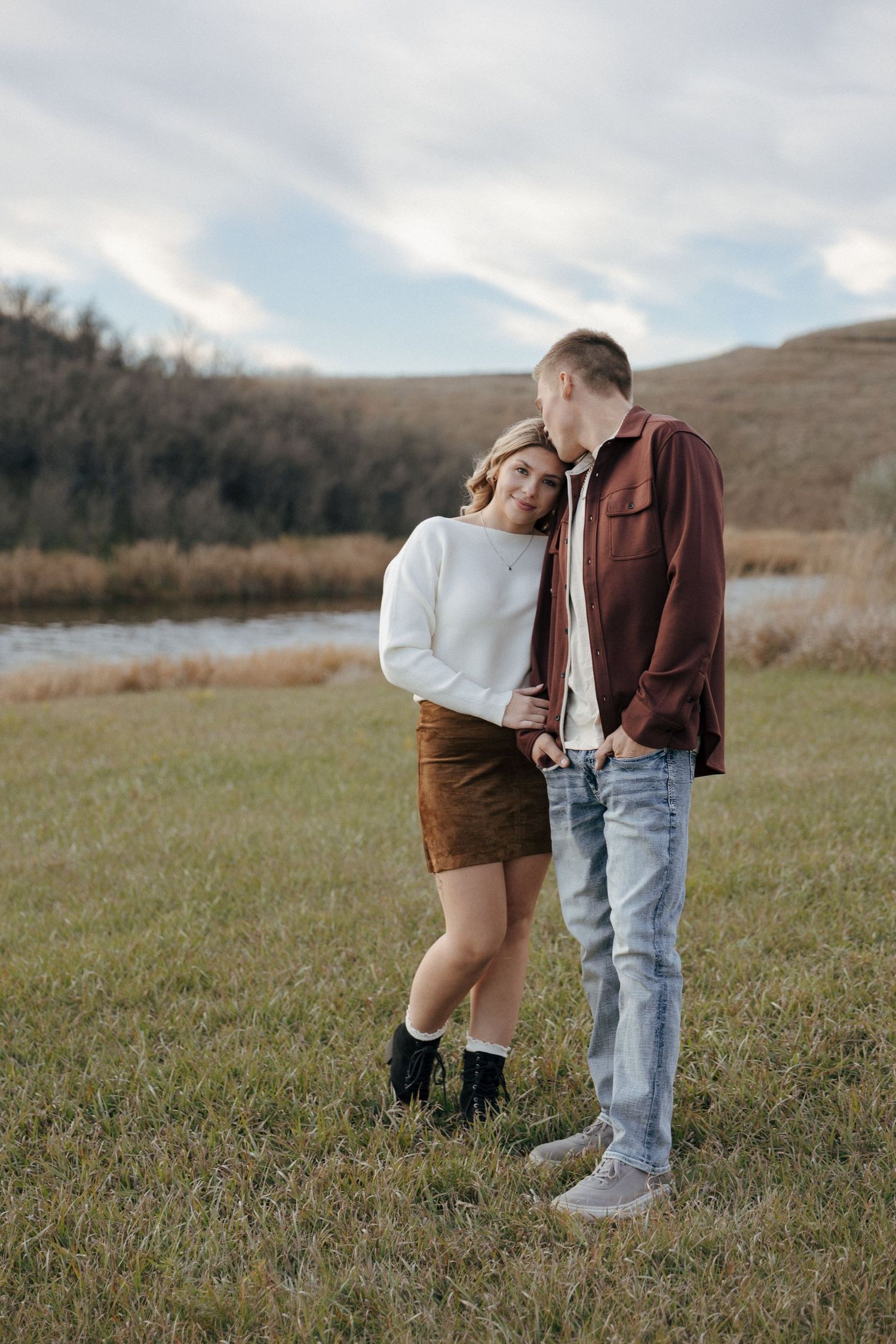 A romantic couple embraces while standing in a meadow with mountains and cloudy skies in the distance.
