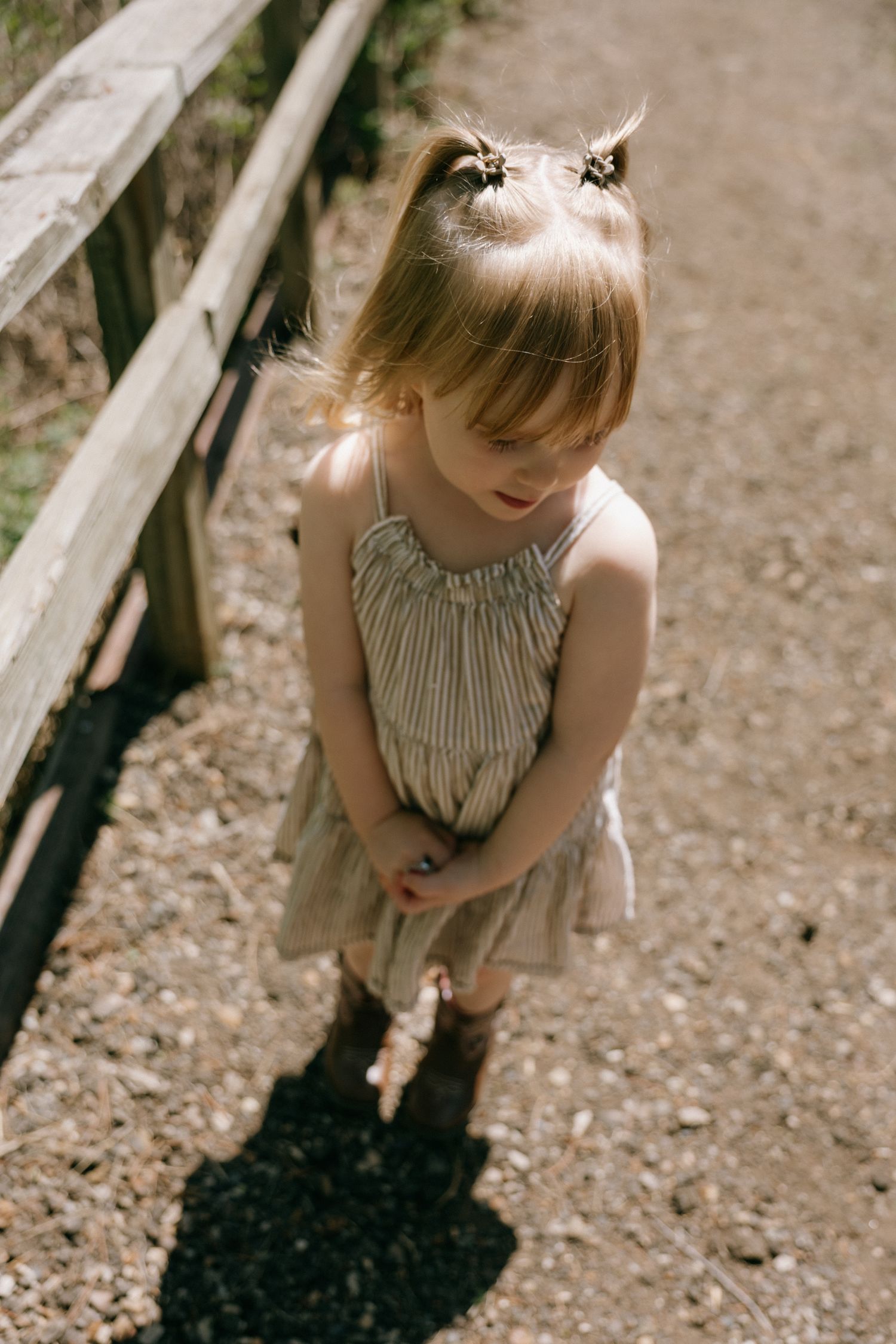 Young girl in striped sundress and cowboy boots standing on a gravel path beside a wooden fence.