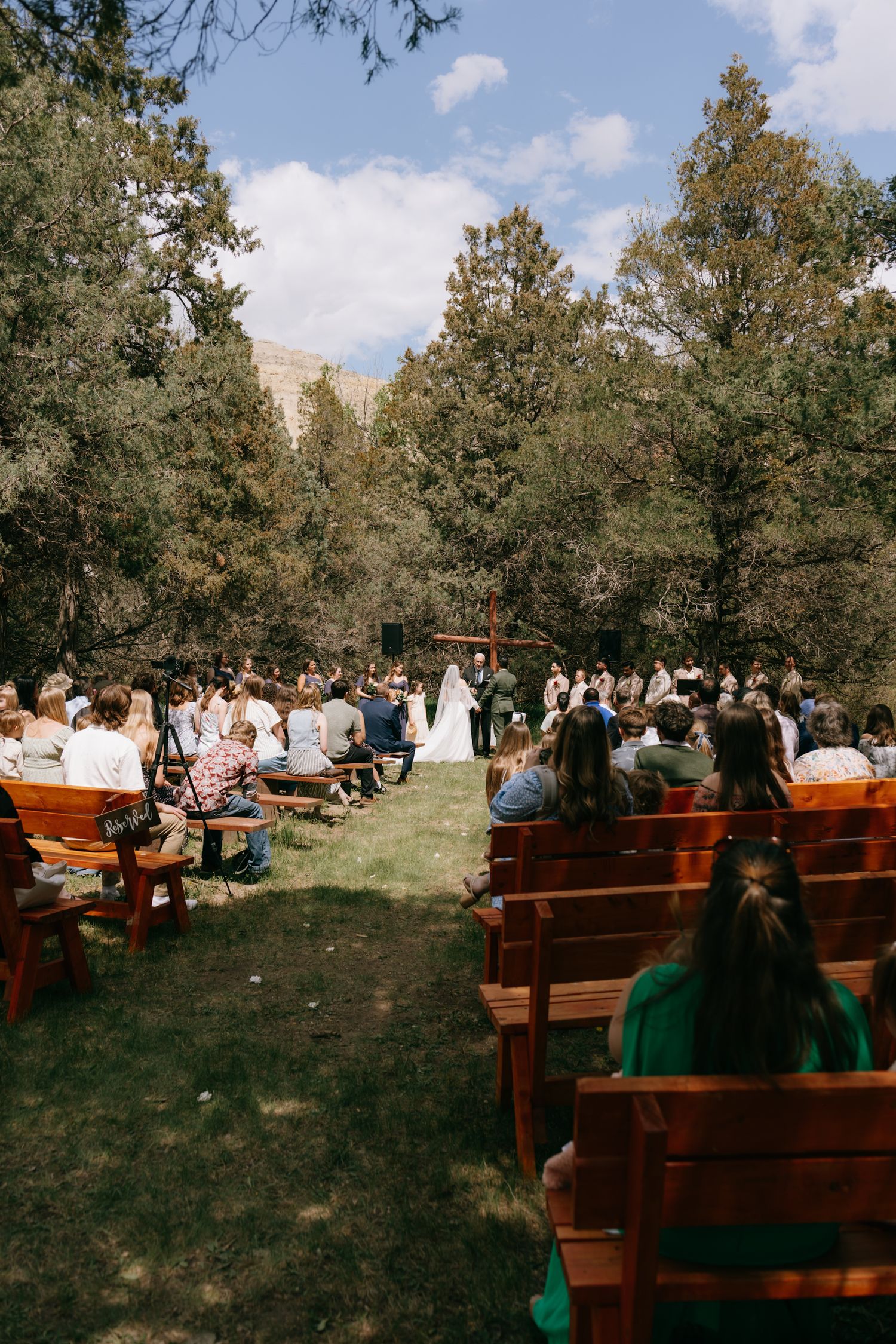 Outdoor wedding ceremony in a forest clearing with guests seated on wooden benches facing the couple at a rustic altar.