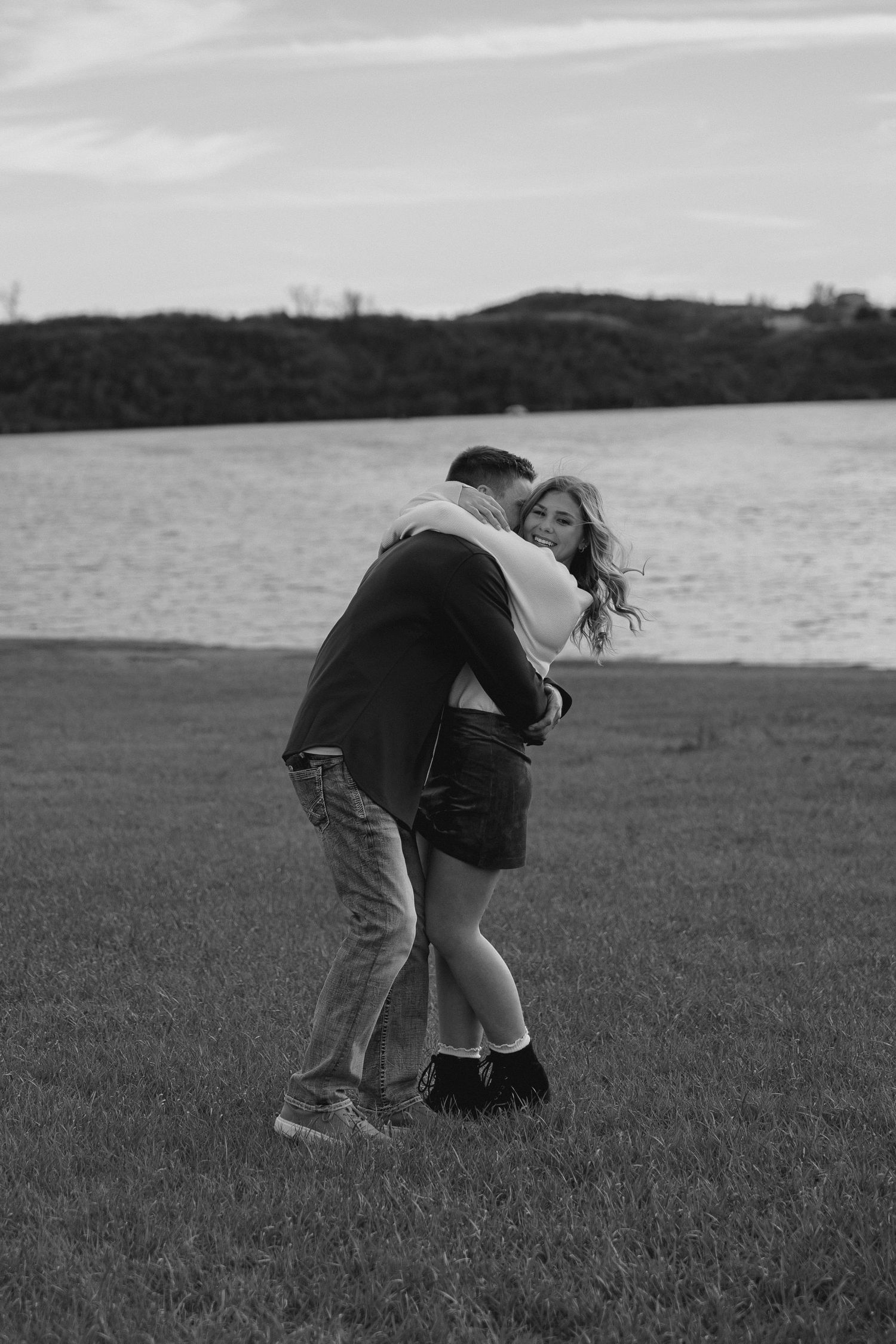 Romantic couple embraces by a lake on grassy shore in black and white photography.
