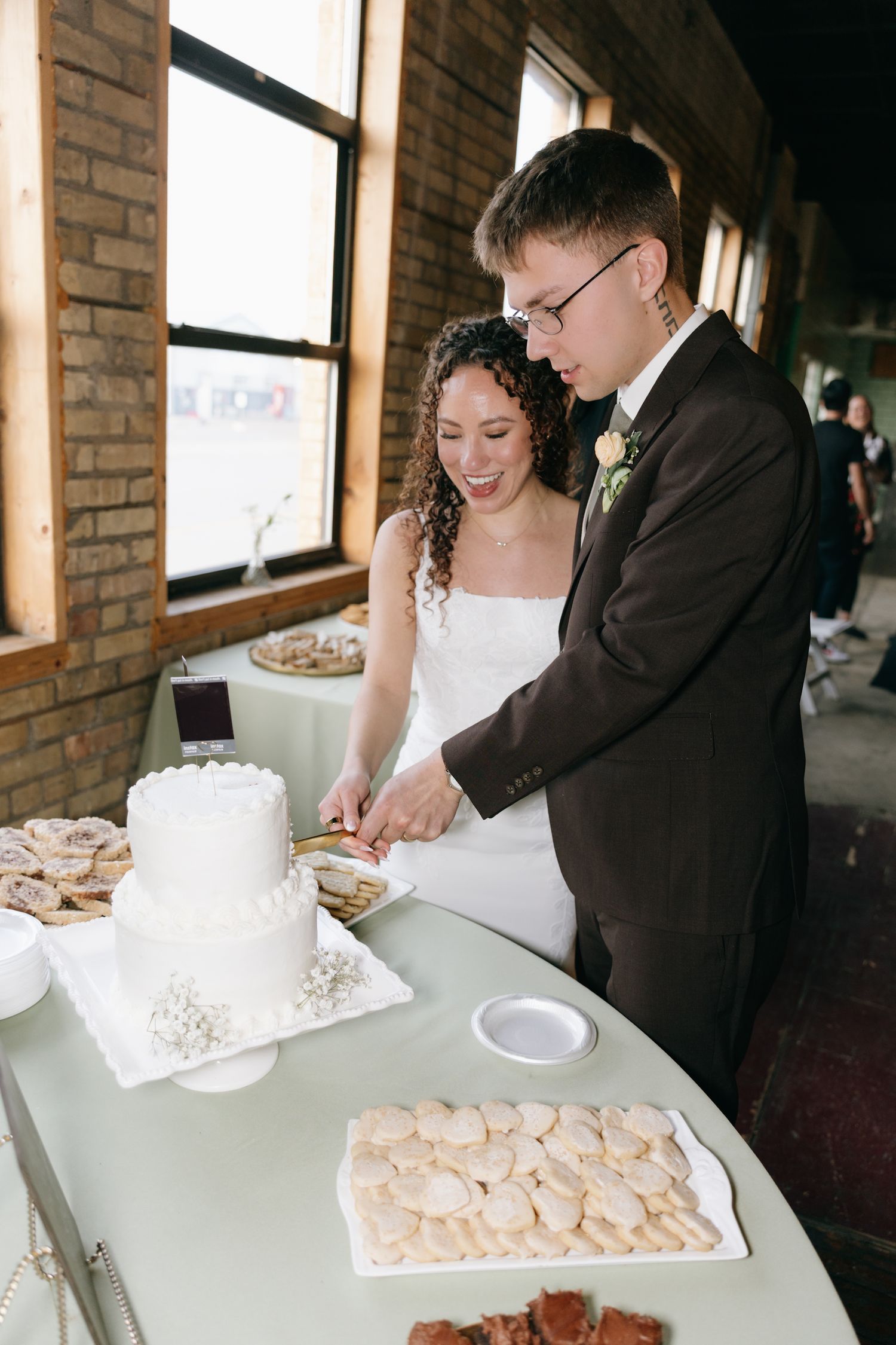 Bride and groom cutting white wedding cake at reception table with assorted cookies displayed nearby.