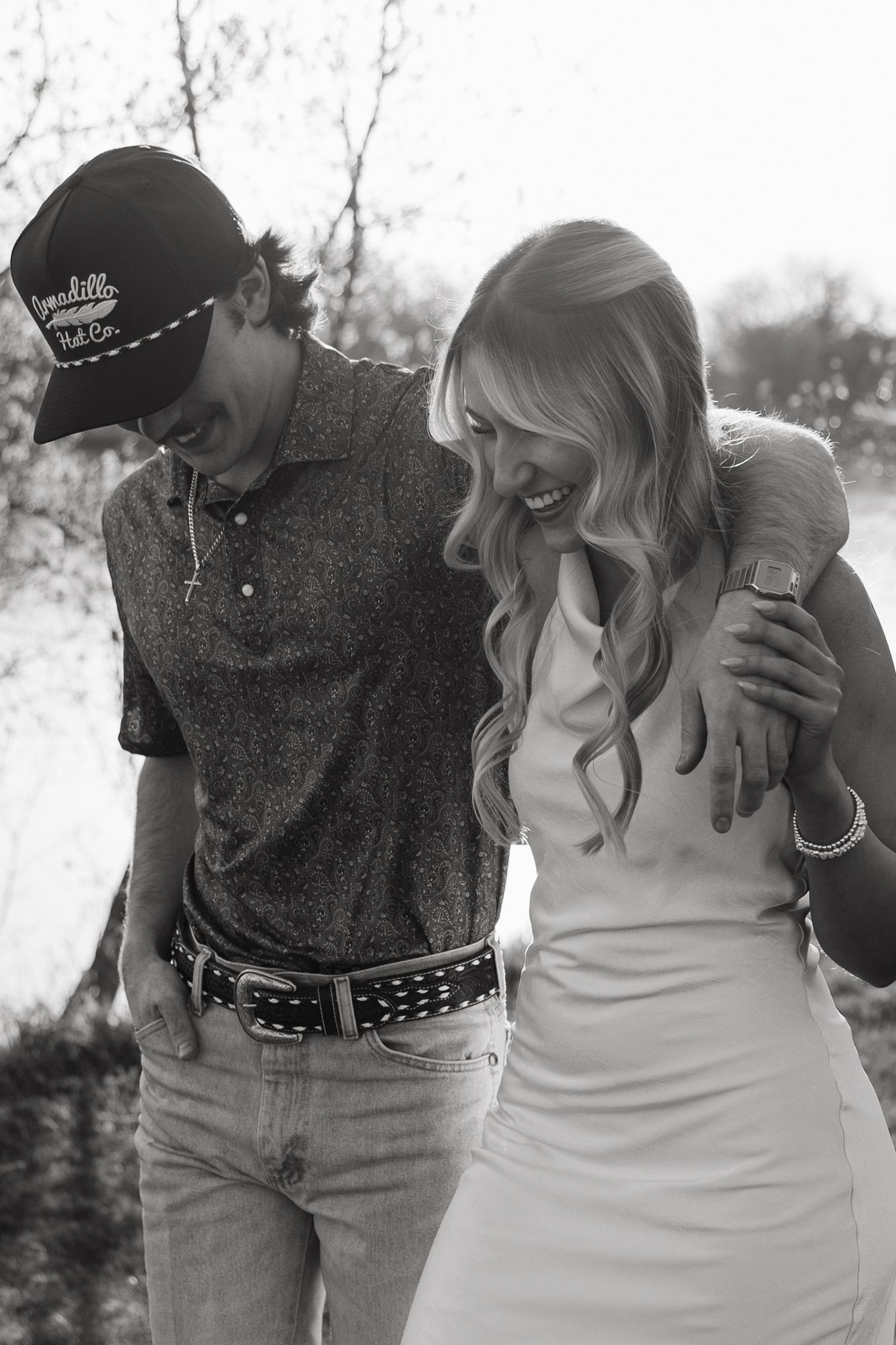 Black and white photo of a laughing couple walking together outdoors, man in cap and polo with arm around woman in white dress.