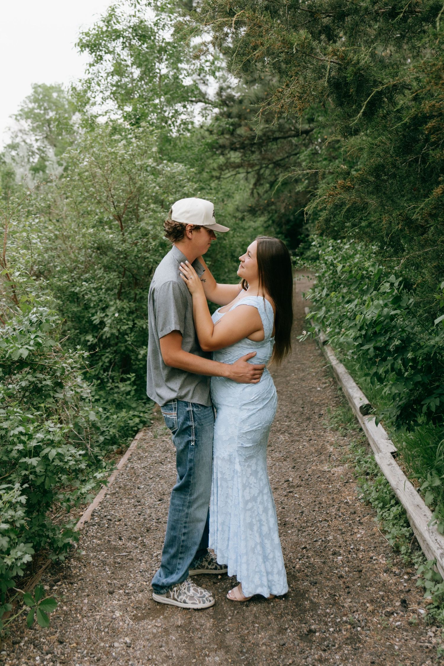A couple embraces on a tree-lined path, the woman in a blue dress and man in jeans and a cap, sharing a romantic moment.