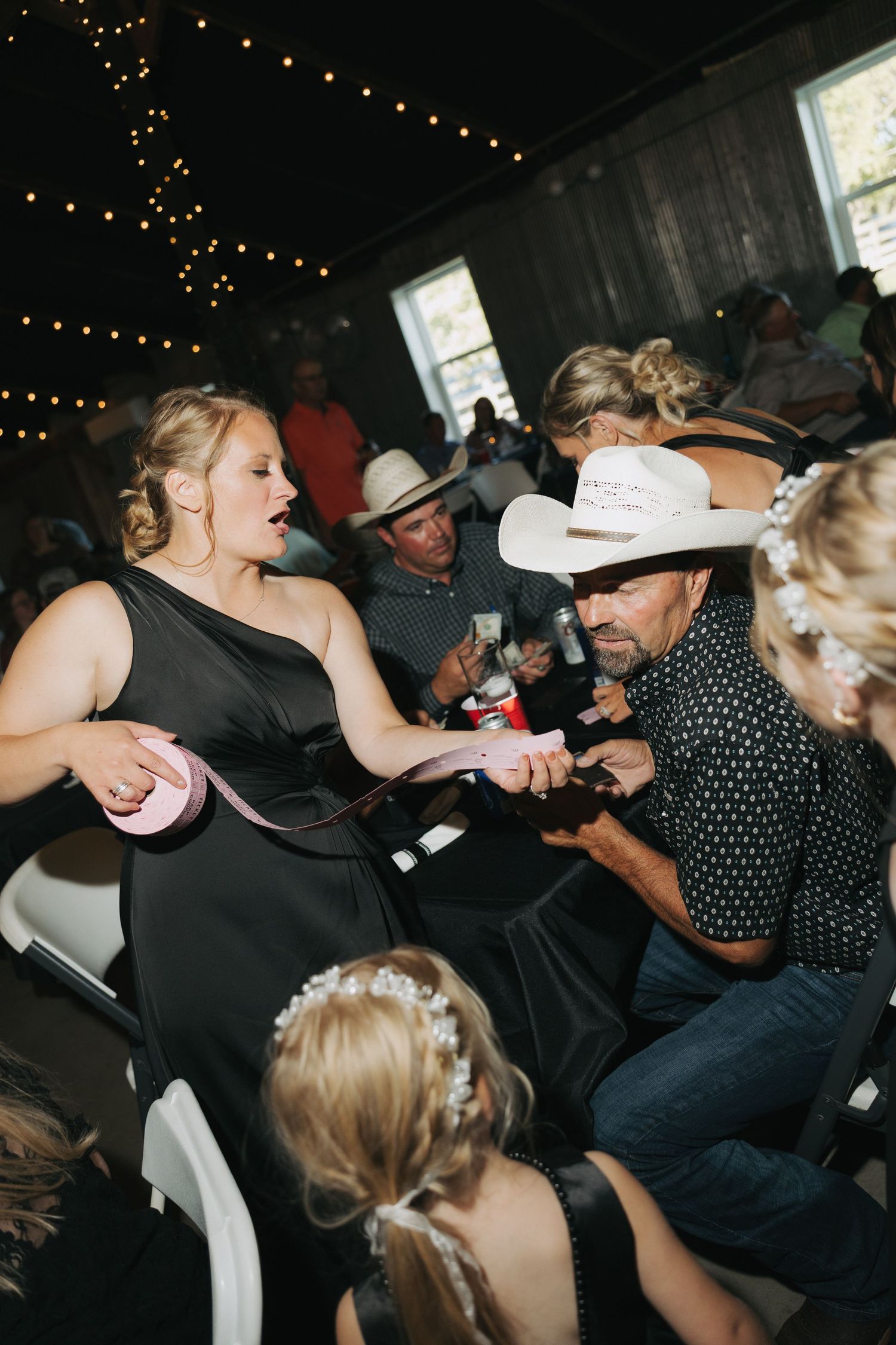 Guests in western attire interact at a rustic wedding reception with string lights in a barn venue.