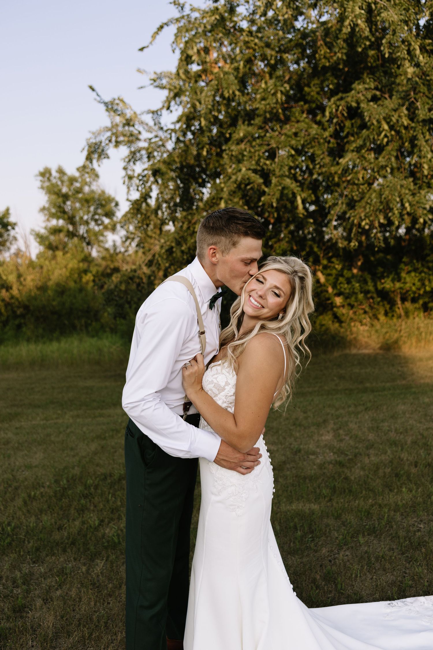 A groom in white shirt and suspenders kisses his smiling bride in a white gown outdoors near lush green trees.