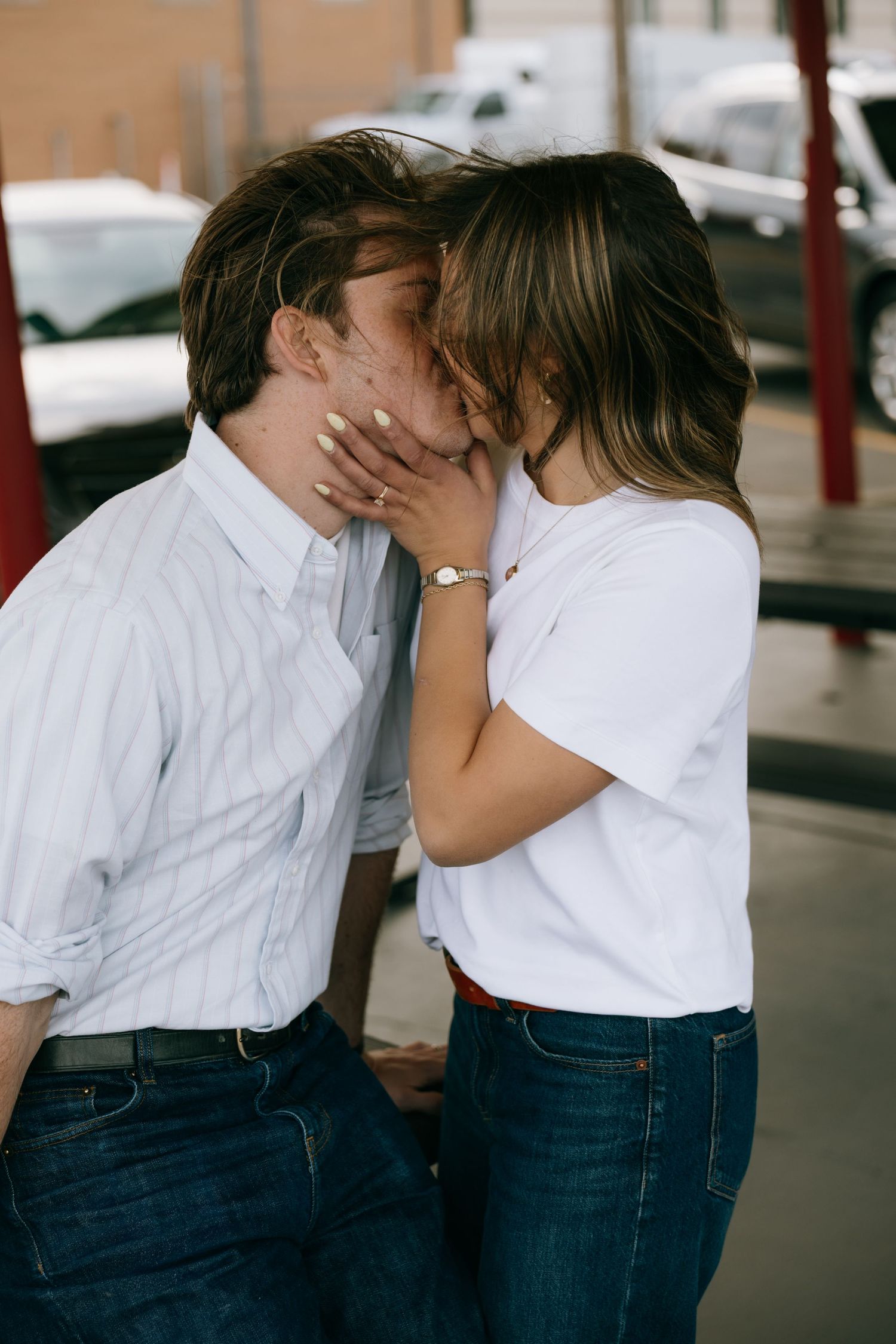A young couple sharing a romantic kiss outdoors, dressed casually in jeans and button-up shirts near a parking lot.