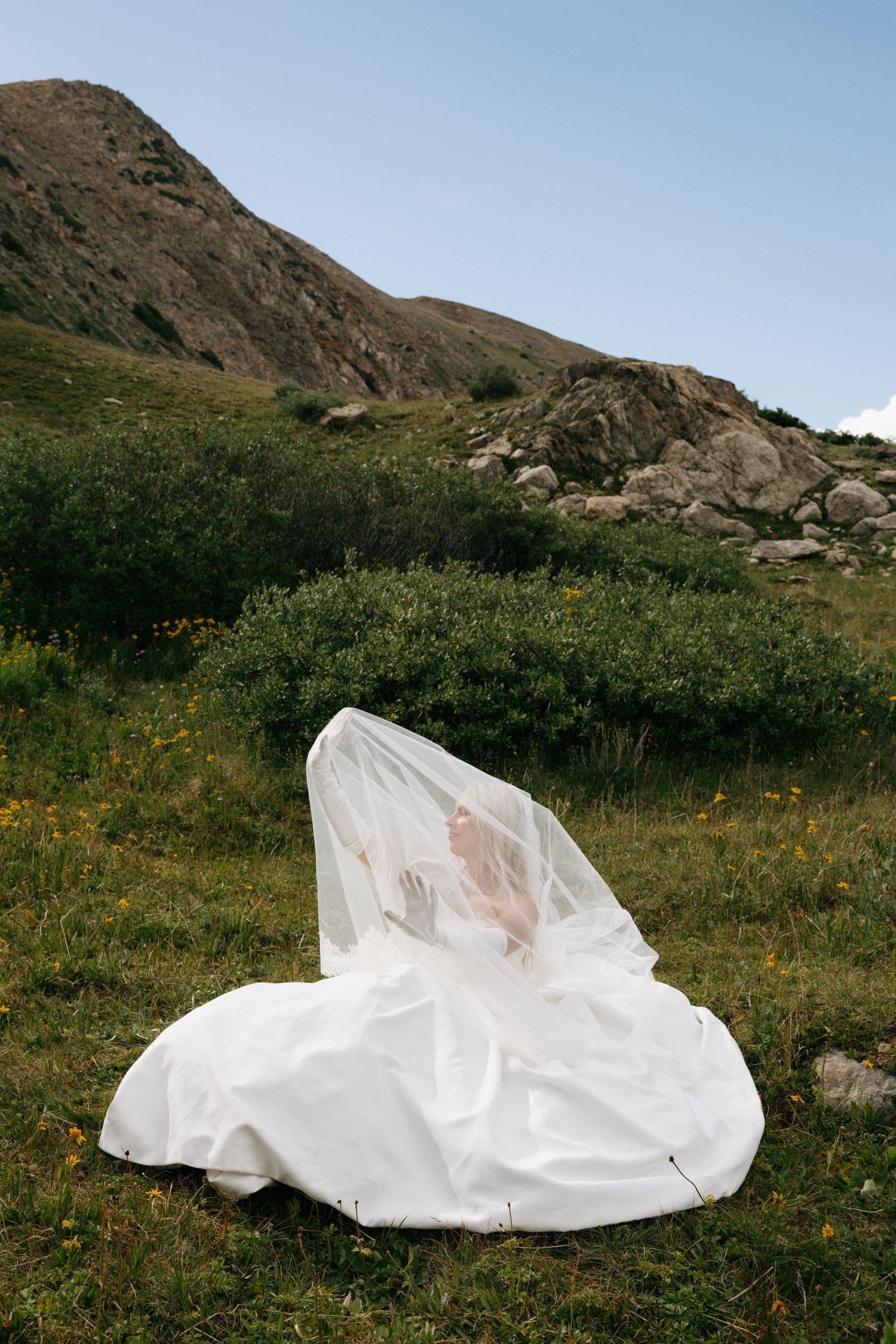 Bride in white wedding dress and veil sitting in alpine meadow with rocky mountain landscape and blue sky.