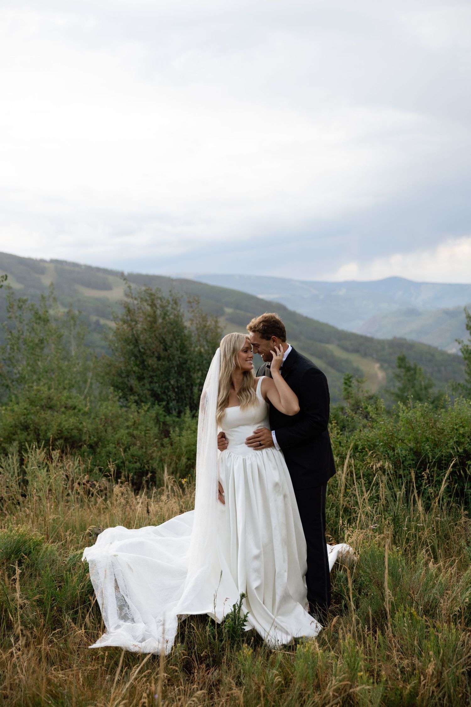 Bride and groom sharing a romantic moment in a mountain meadow, with sweeping hills and cloudy skies behind them.