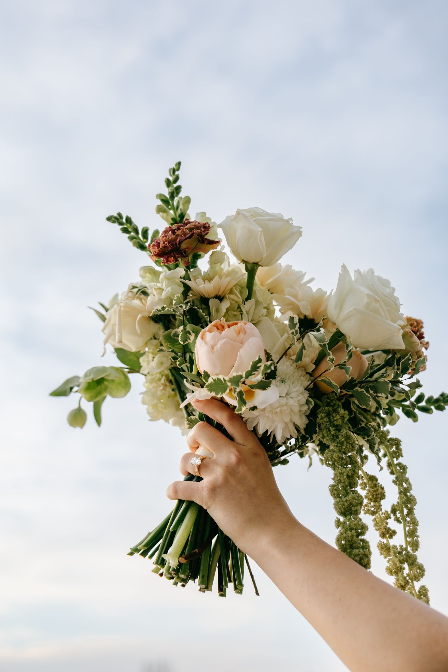 Hand holding a bridal bouquet of white roses, peonies, and greenery against a soft blue sky.