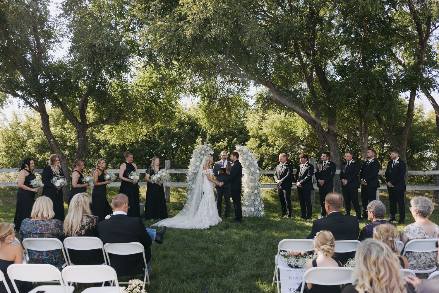 Outdoor wedding ceremony under lush trees with bridal party in black attire and guests seated in white chairs.