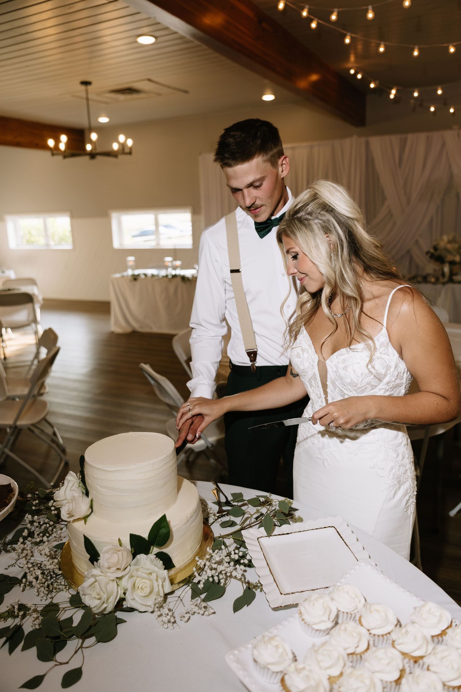 Bride and groom cutting white wedding cake decorated with roses at elegant reception venue.