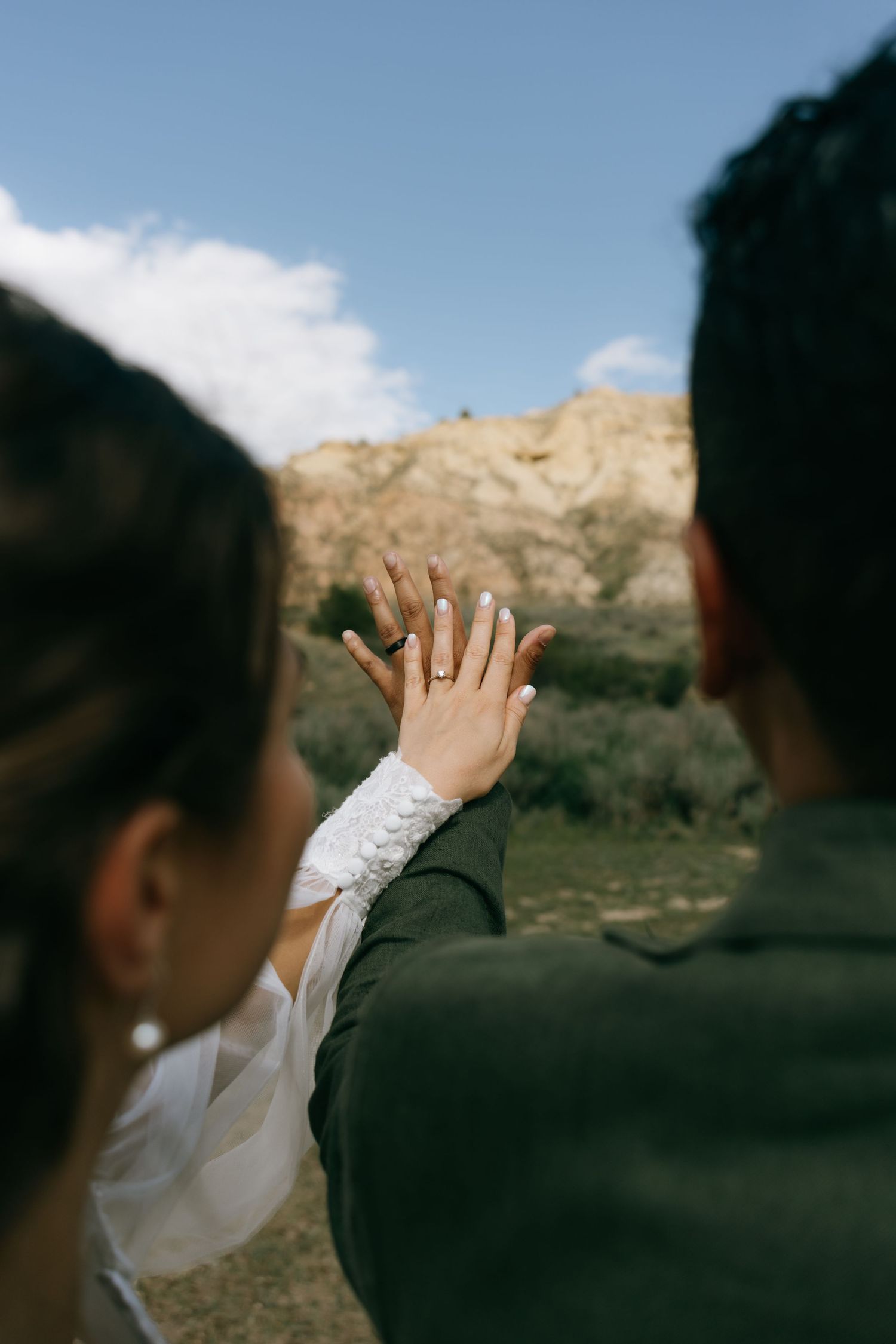 Newlywed couple holding hands outdoors, showcasing wedding ring against scenic mountain backdrop.