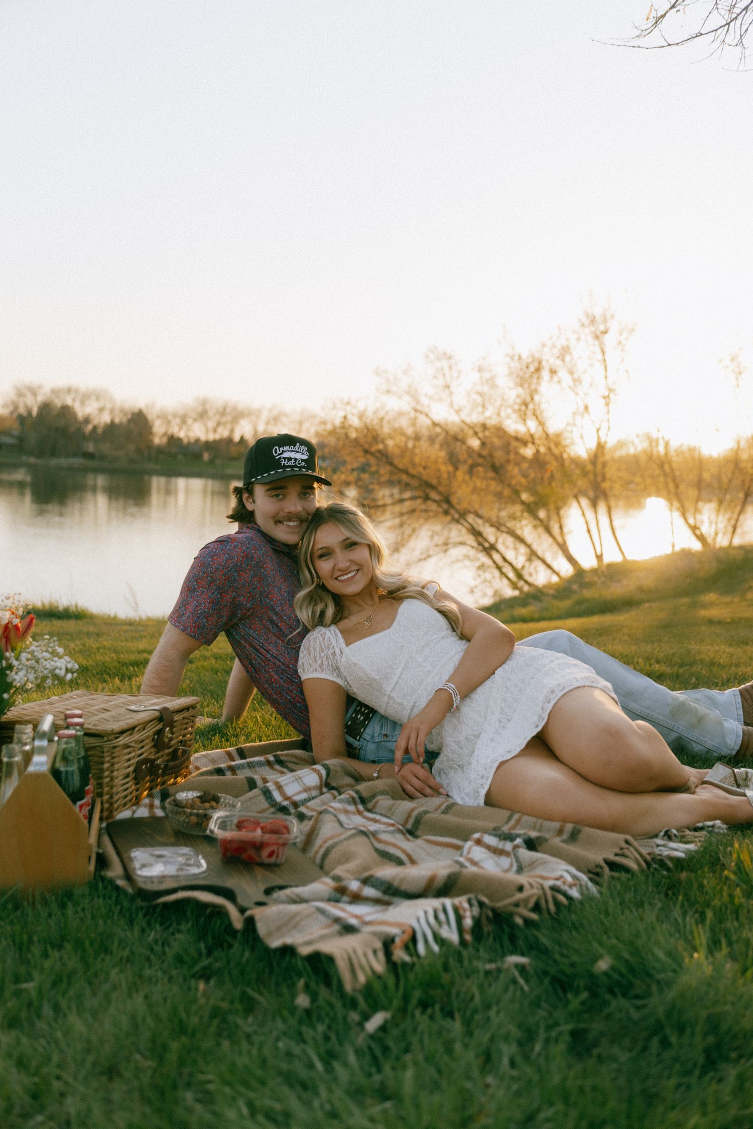 Couple relaxing on a plaid blanket during a romantic riverside picnic at golden hour sunset.