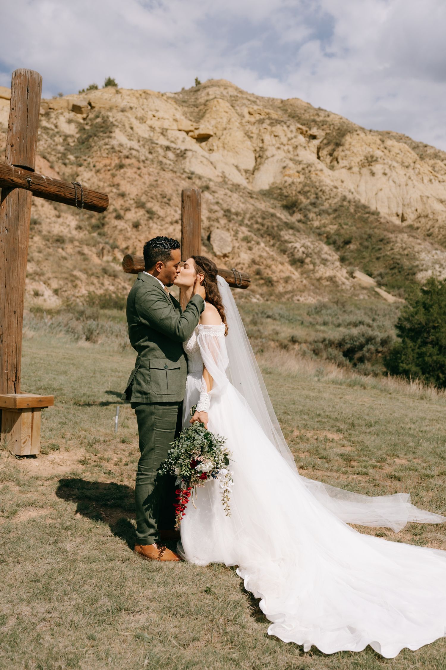 Bride and groom share first kiss at outdoor wedding ceremony with wooden cross and scenic rocky hills backdrop.