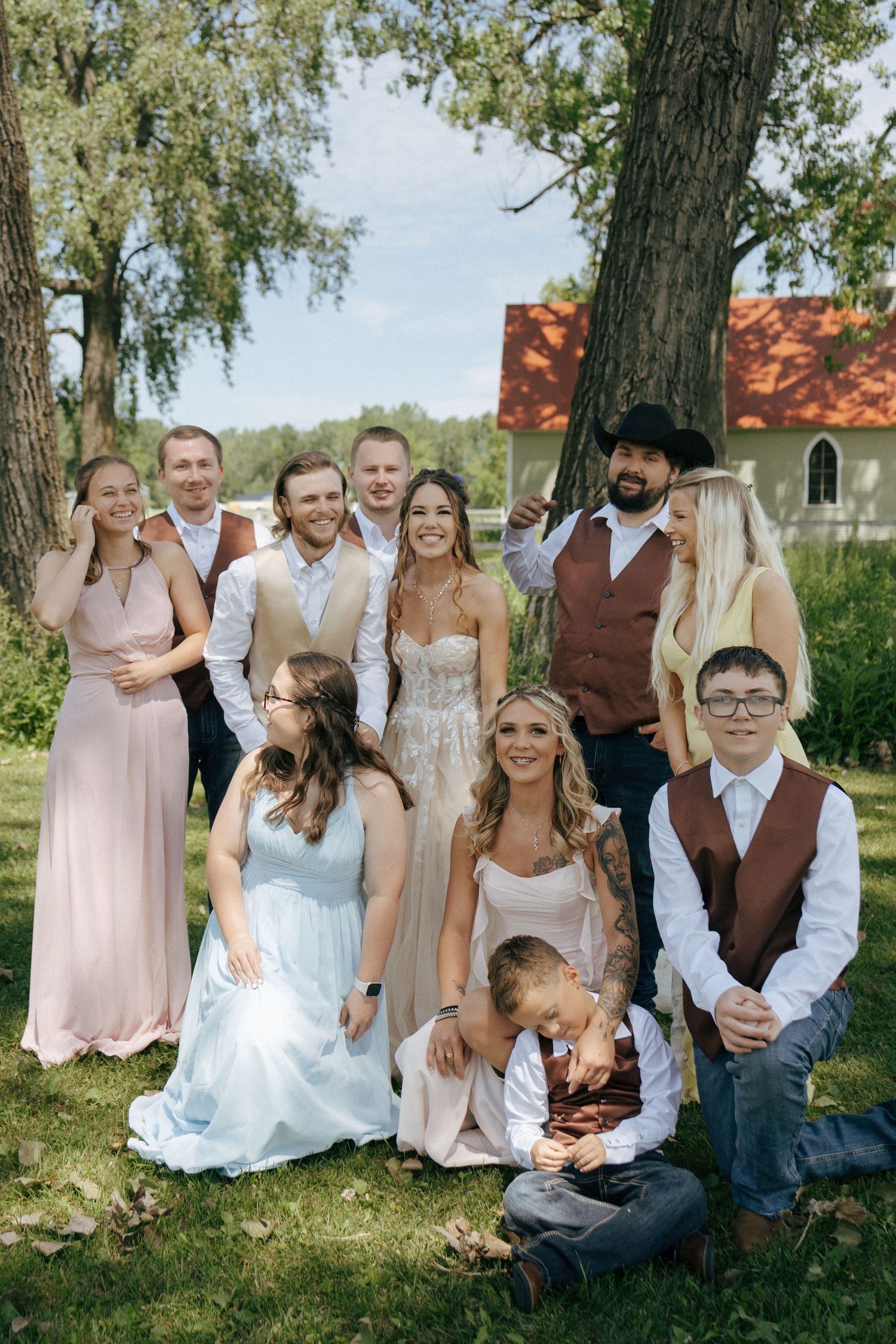 Wedding party poses outdoors in casual country attire with pink bridesmaid dresses and brown vests.