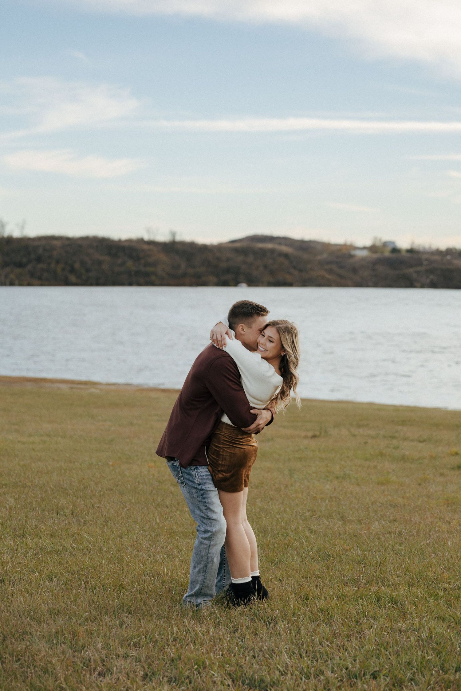 Two people share a romantic moment in a lakeside field during sunset.