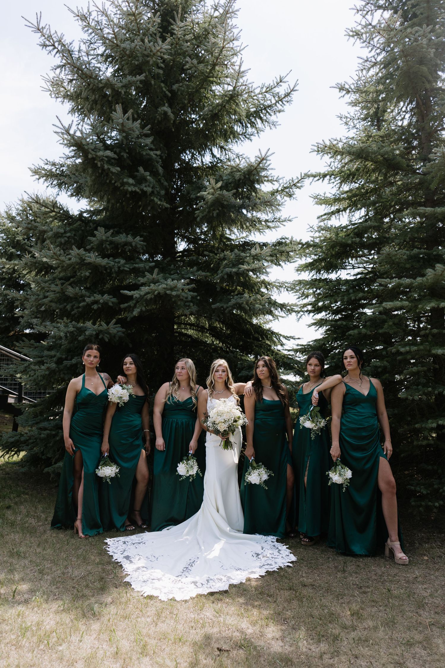 Bride in white gown with six bridesmaids in emerald green satin dresses holding white bouquets before tall pine trees.