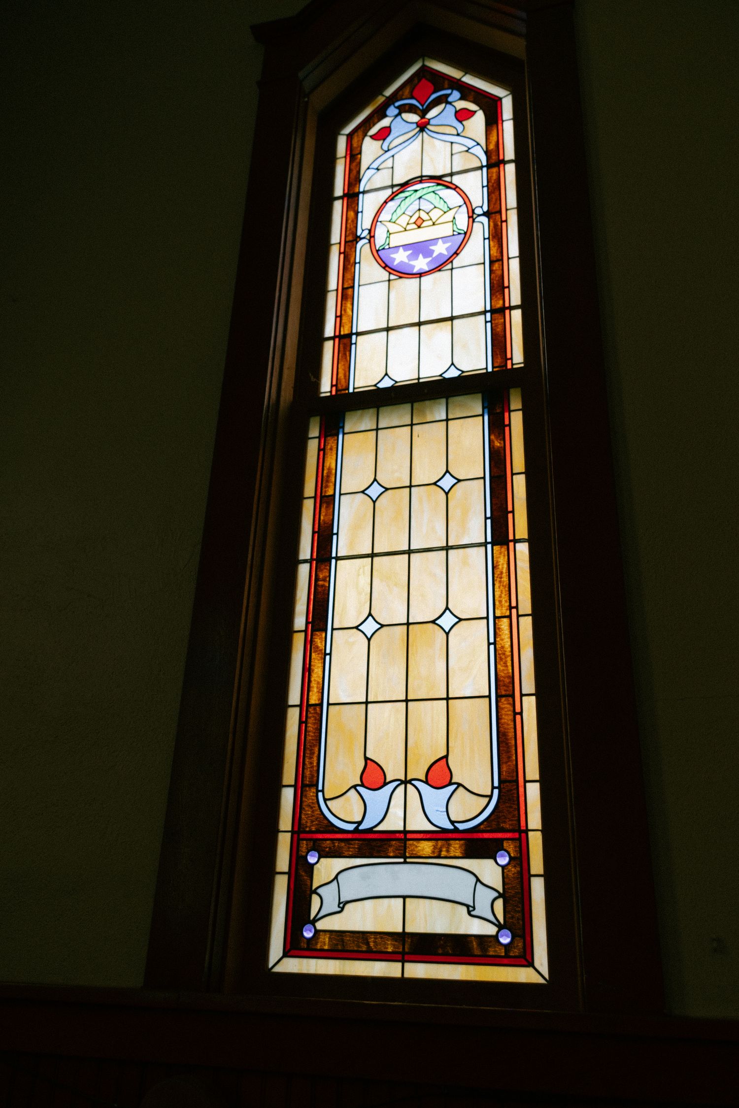 Tall stained glass church window with colorful geometric patterns, floral motifs, and decorative border in wooden frame.