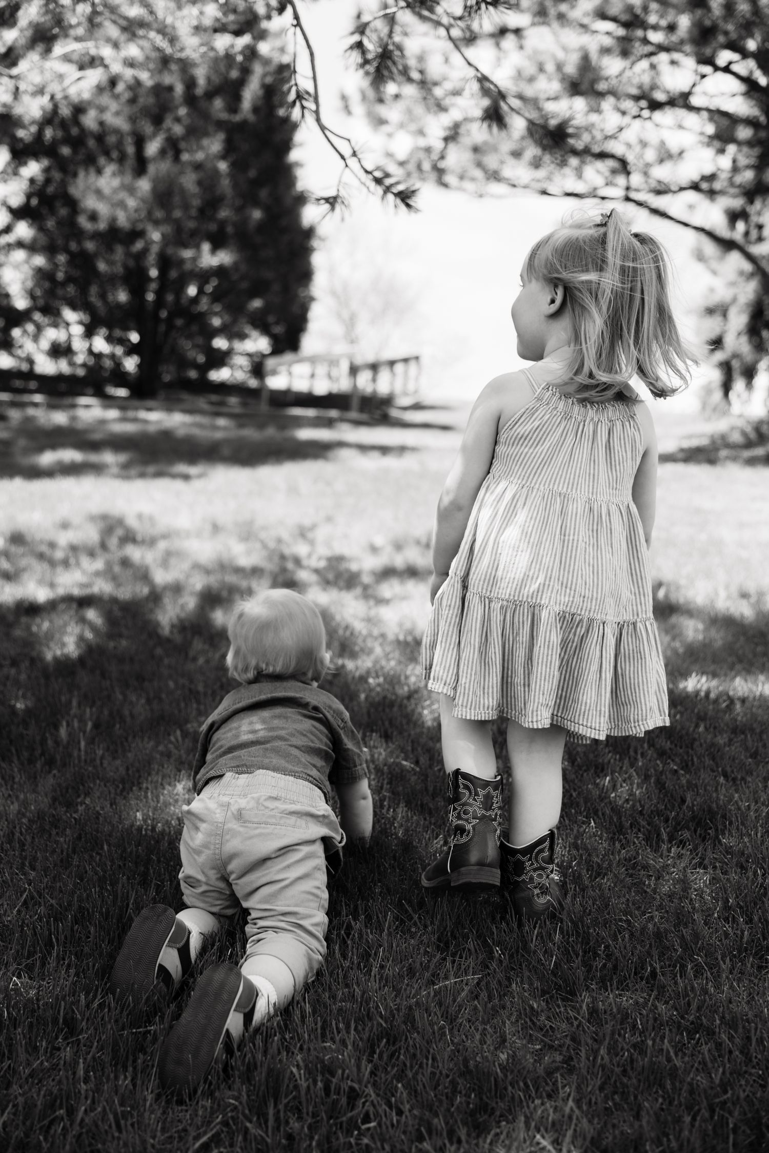 Black and white photo of two young children playing outdoors, a toddler crawling and a girl in a dress standing on grass.