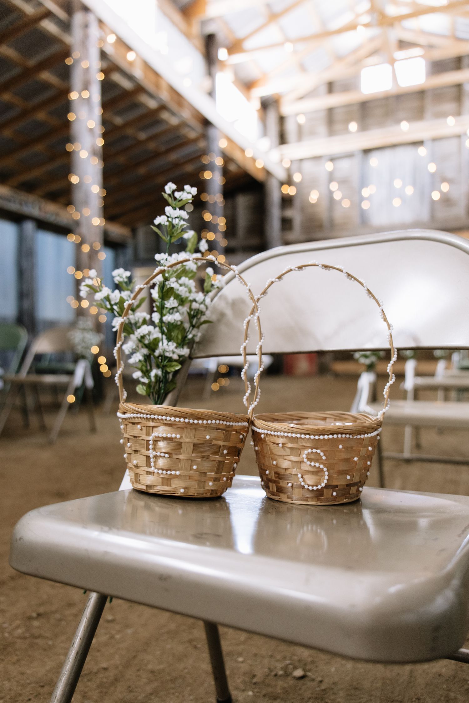 Two wicker flower girl baskets with pearl trim on a metal chair at a rustic barn wedding venue with fairy lights.