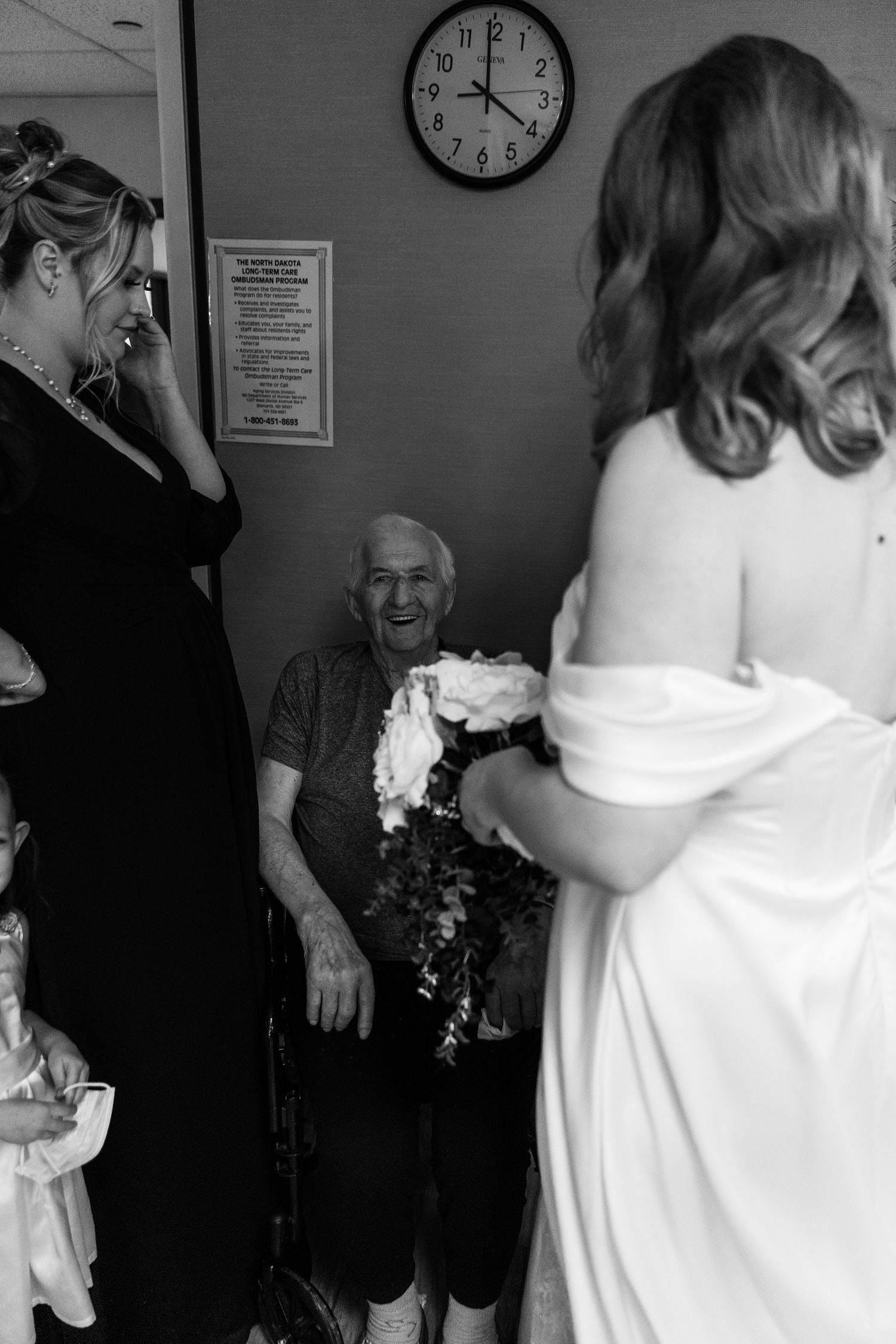 Black and white photo of a bride in white gown showing bouquet to elderly man in wheelchair, emotional wedding moment.