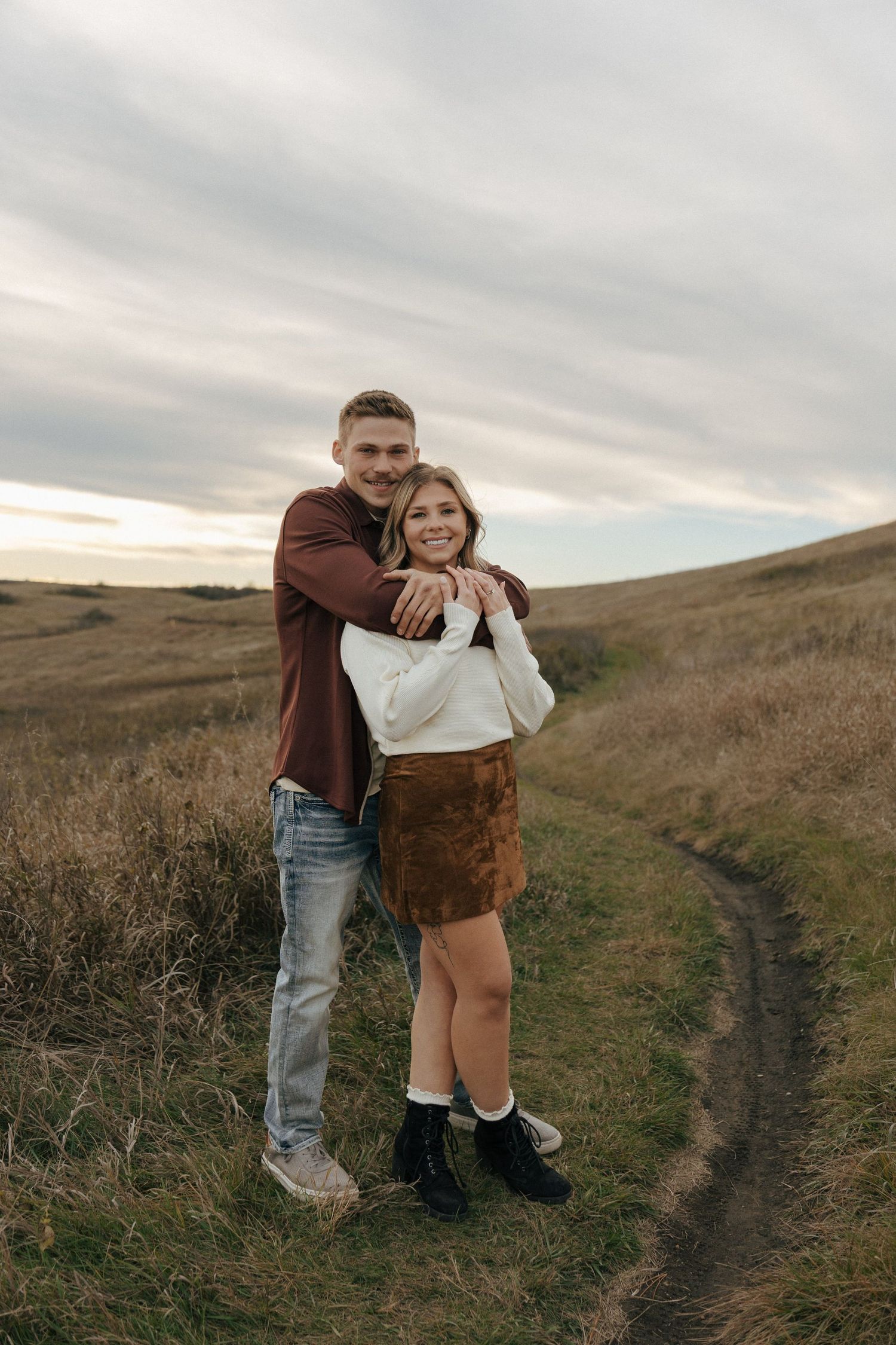 Couple sharing affectionate moment on hiking trail with rolling hills in background.