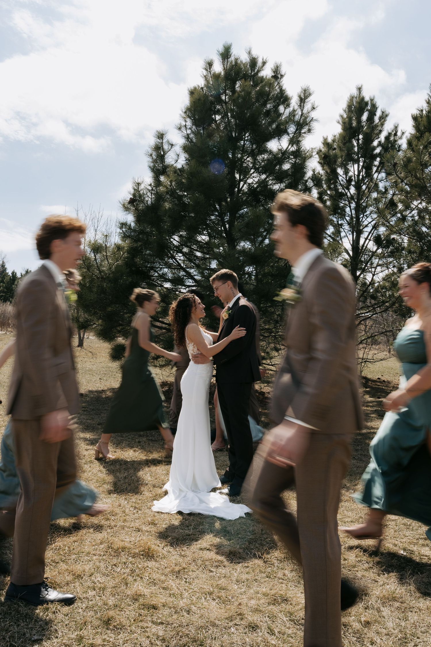 Bride and groom share a romantic moment outdoors as wedding party walks past in brown and teal.