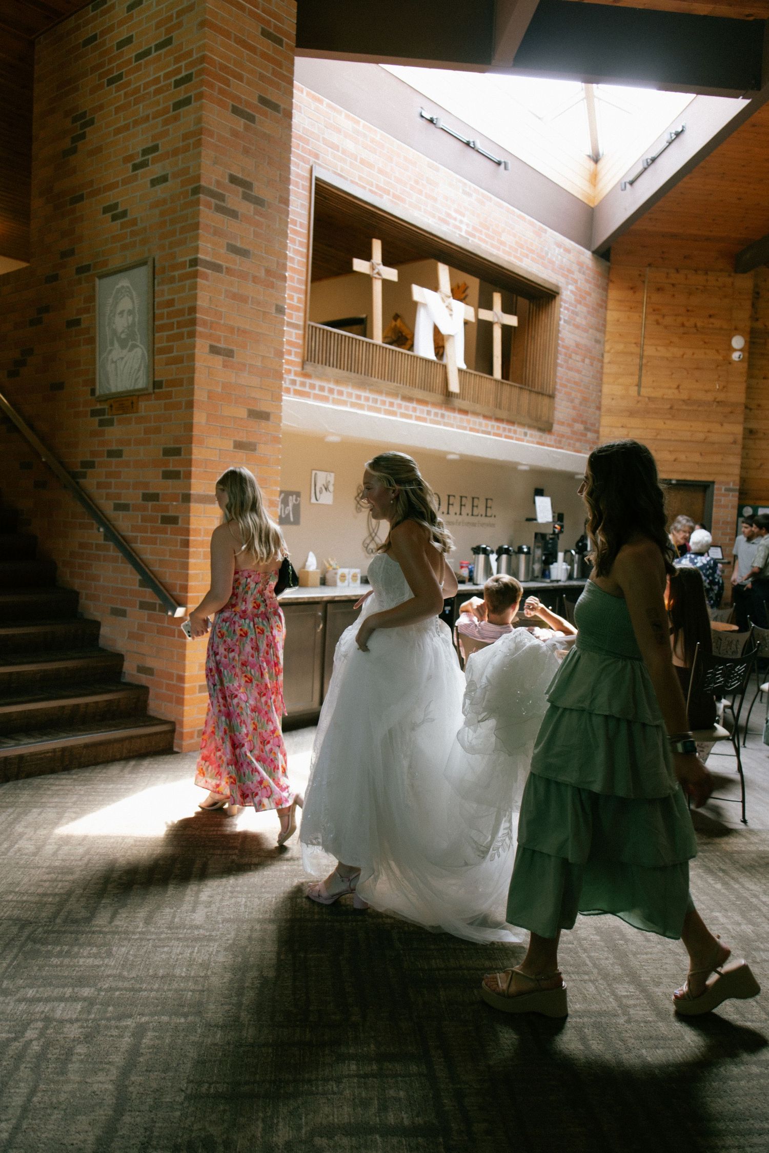 Bride in white gown walks with bridesmaids in floral and green dresses inside a brick church venue.