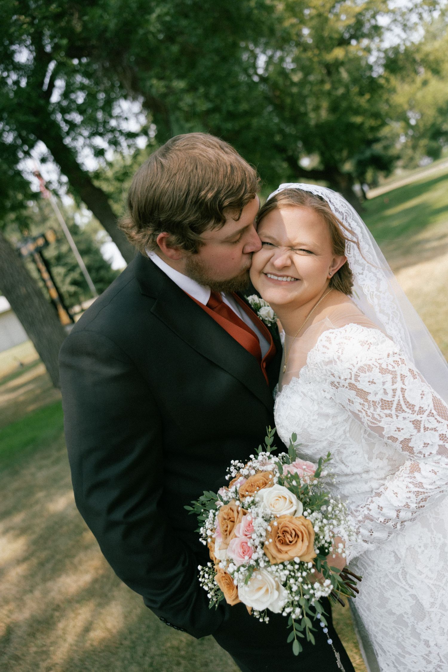 Groom in black suit kisses smiling bride wearing lace dress and veil, holding peach and pink bouquet outdoors.