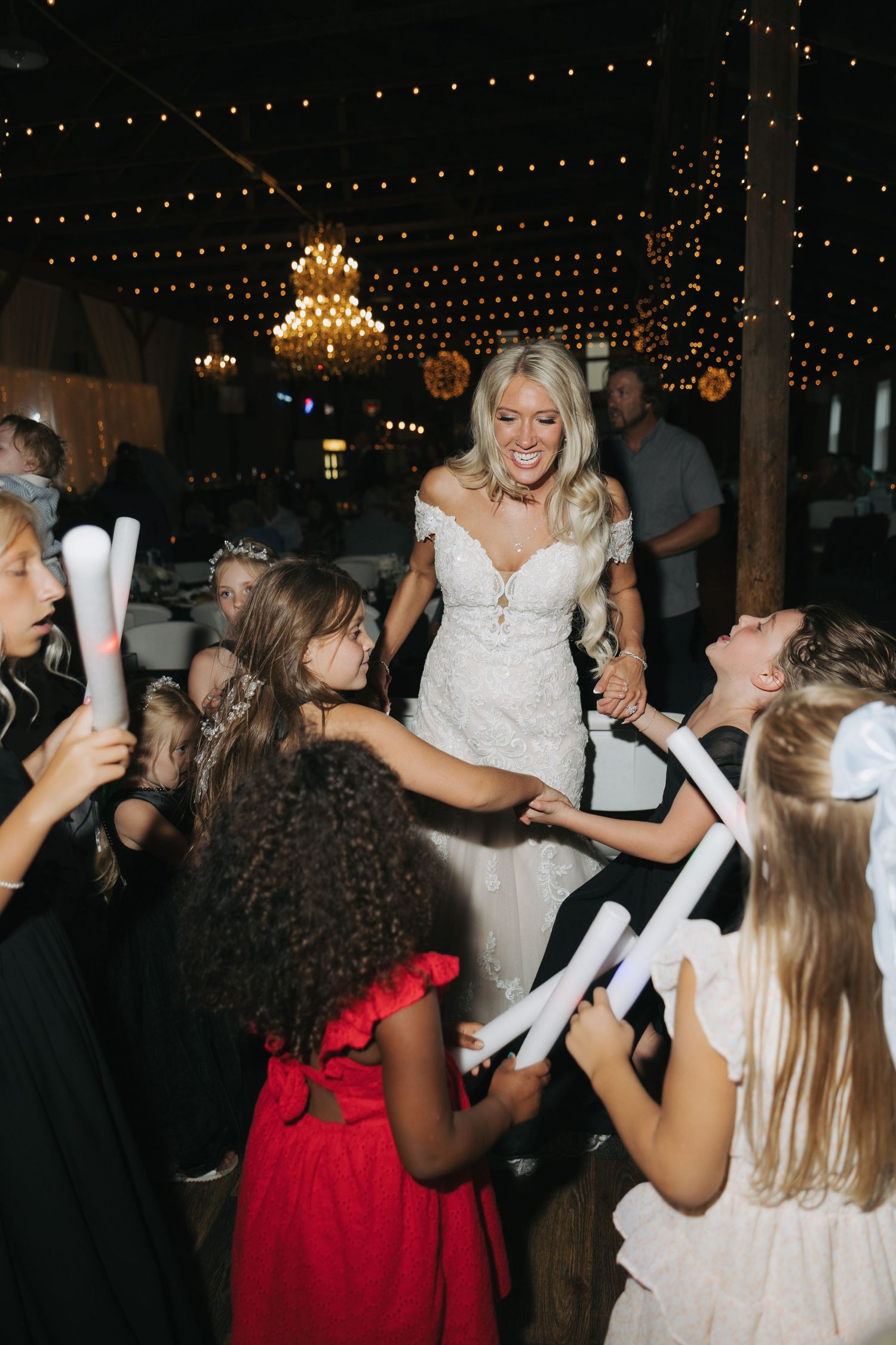 Bride in white lace gown dancing with children holding glow sticks at a wedding reception with string lights.